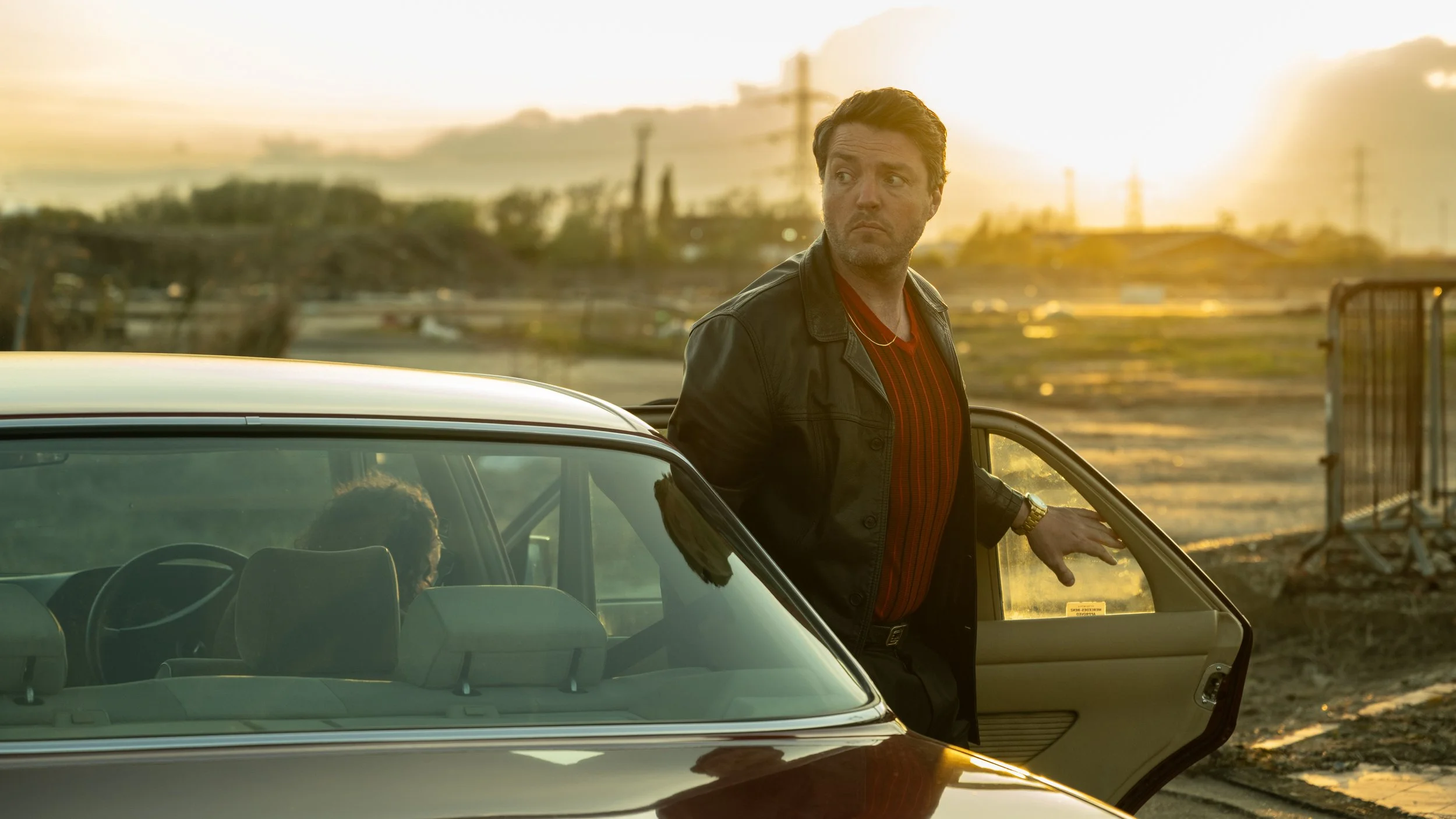 Tom Burke as customs officer Guy Stanton in Legends for Netflix. Tom is pictured at sunset exiting a vintage car in a desolate wasteland wearing a red t shirt and leather jacket. Photo by Sally Mais