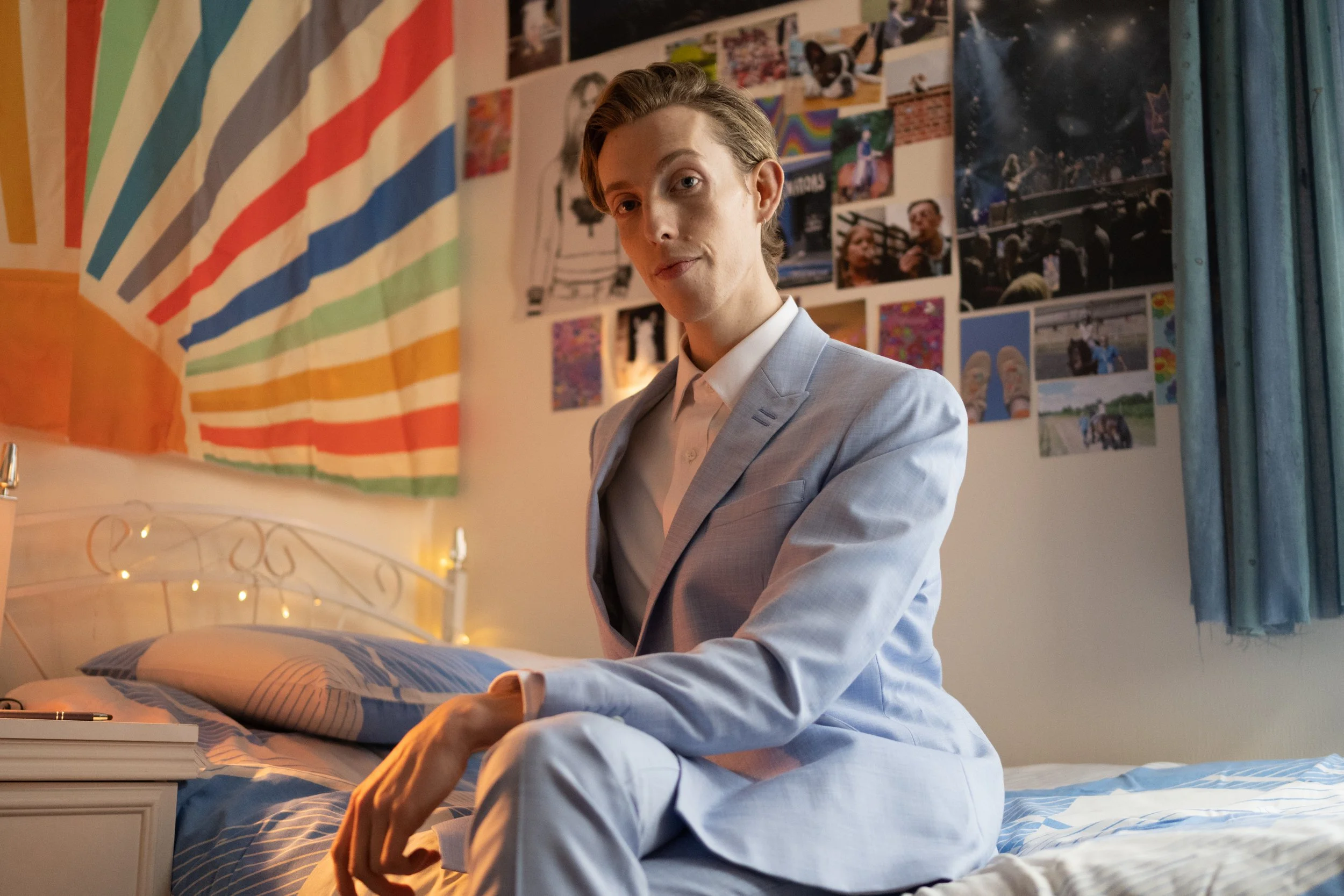 Harry Trevaldywn as his character Billie, sits on a bed in a small bedroom with posters on the wall 