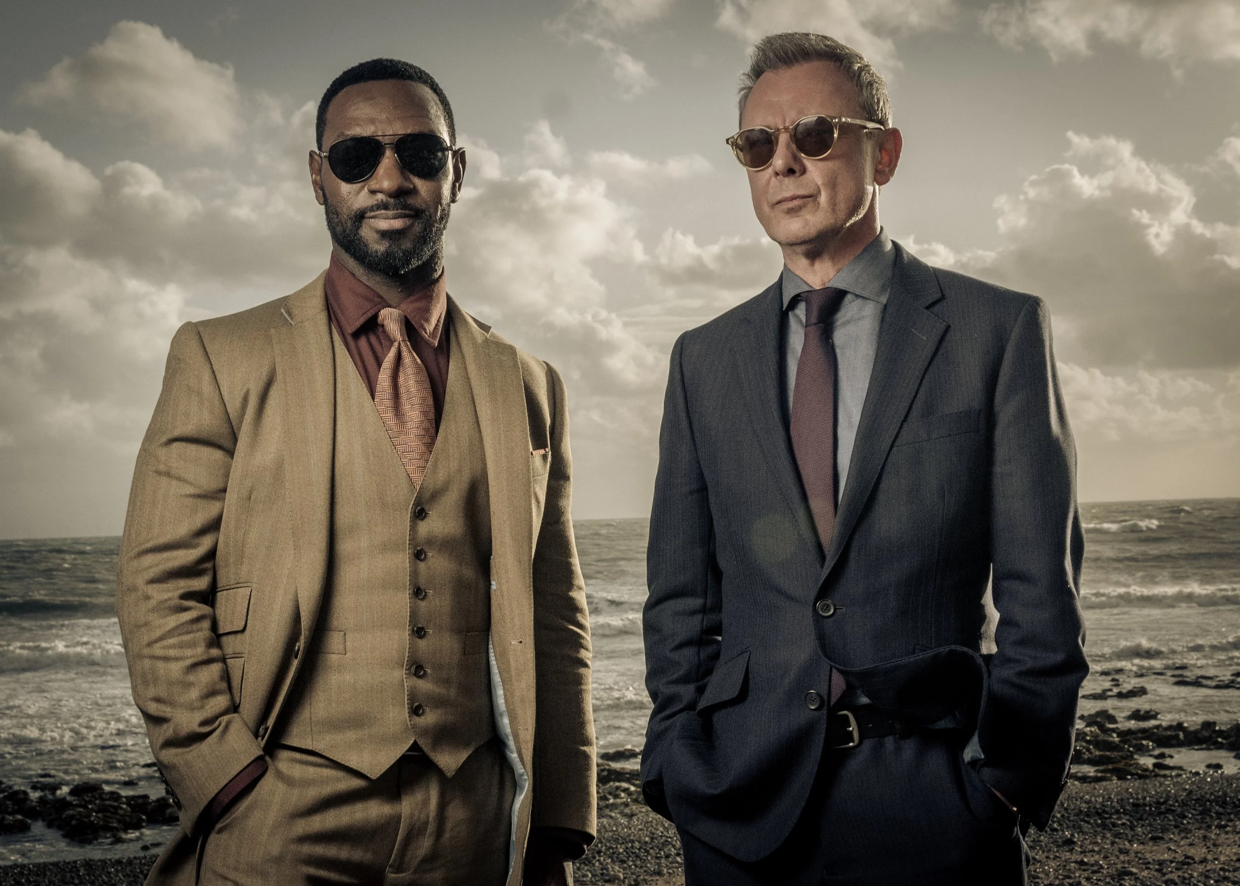 Richie Campbell as Branson and John Simm as Grace in the dectective series Grace for ITV. They stand on a beach in smart suits with sunglasses on on  Photo by series photographer Sally Mais who shoots both stills and key art for the series 