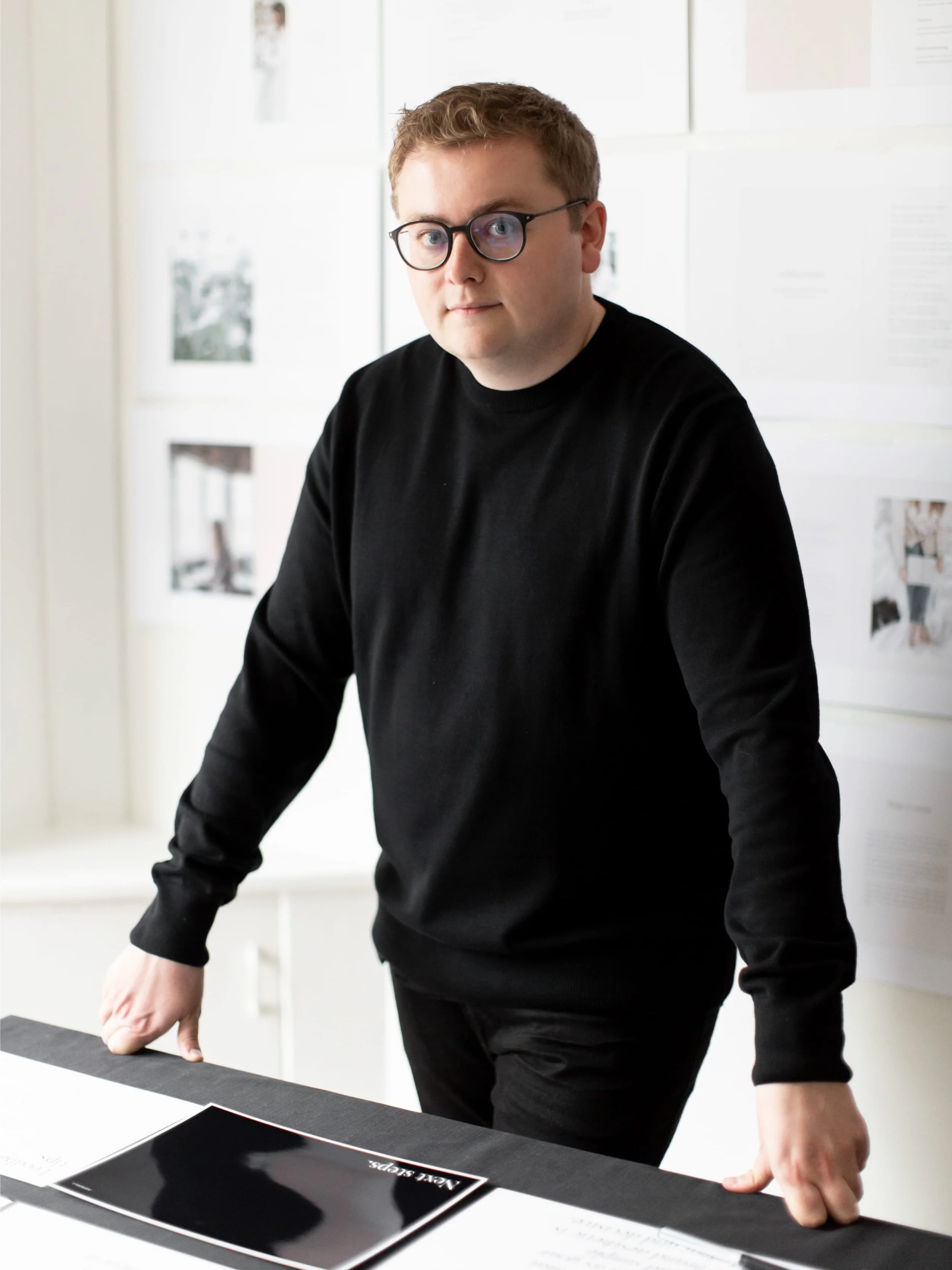 Jack Watkins standing in his light-filled studio, leaning on a desk covered in brand design materials