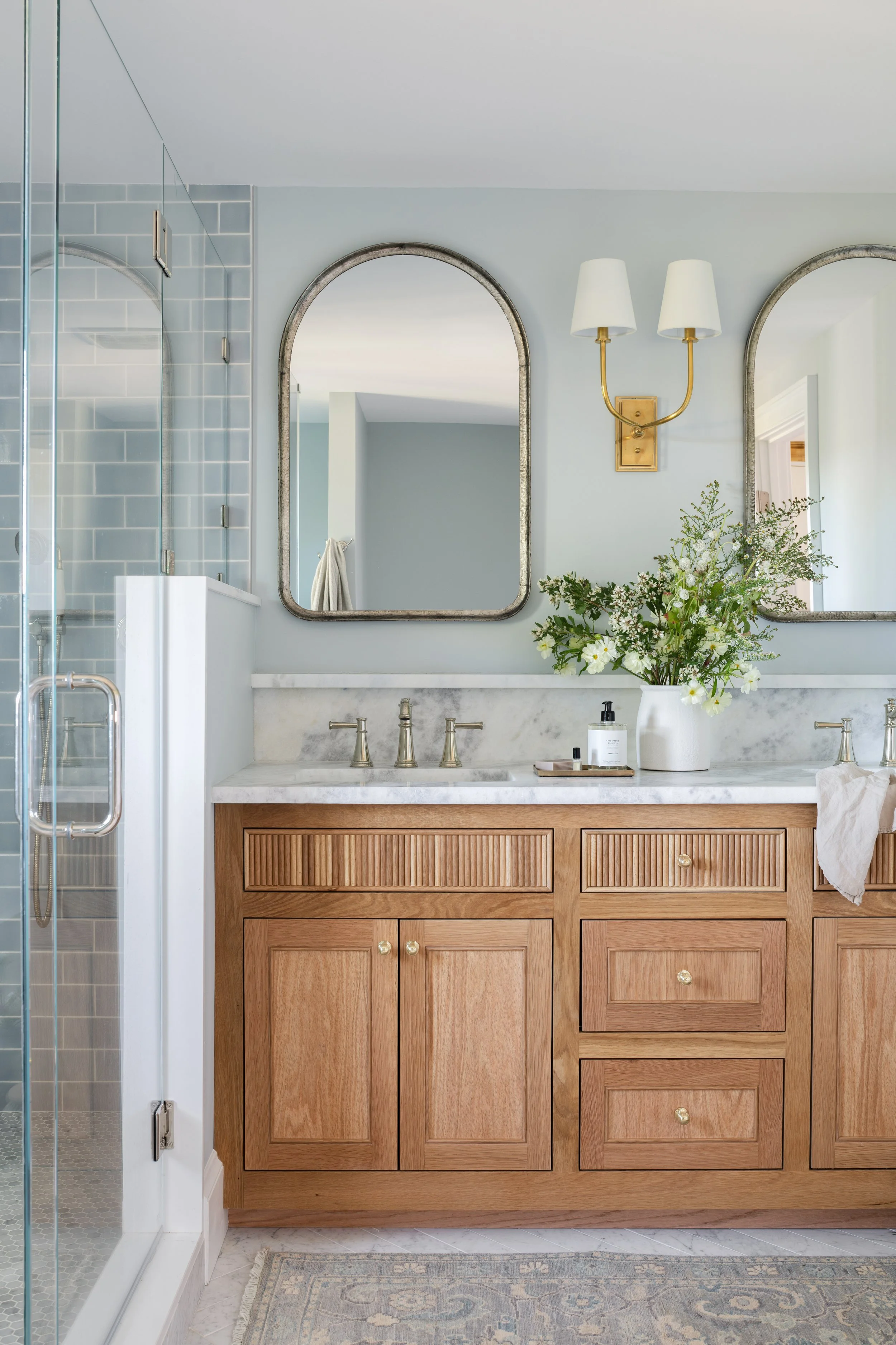 Sustainable Cape Cod home design shines in this bathroom with a wooden vanity, marble countertop, two mirrors, a bouquet of flowers, a wall sconce, and a shower with blue tile walls.