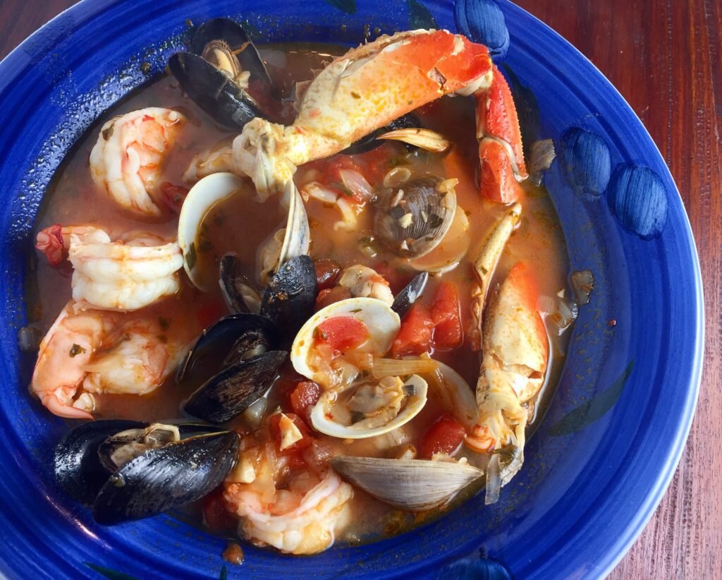Seafood bowl with shrimp, mussels, crab claws, and clams in a tomato-based broth on a blue plate.