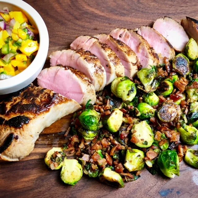 Sliced seared pork tenderloin, sautéed Brussels sprouts with chopped bacon, a serving of fruit salsa with mango, red onion, and cucumber, all on a wooden cutting board.