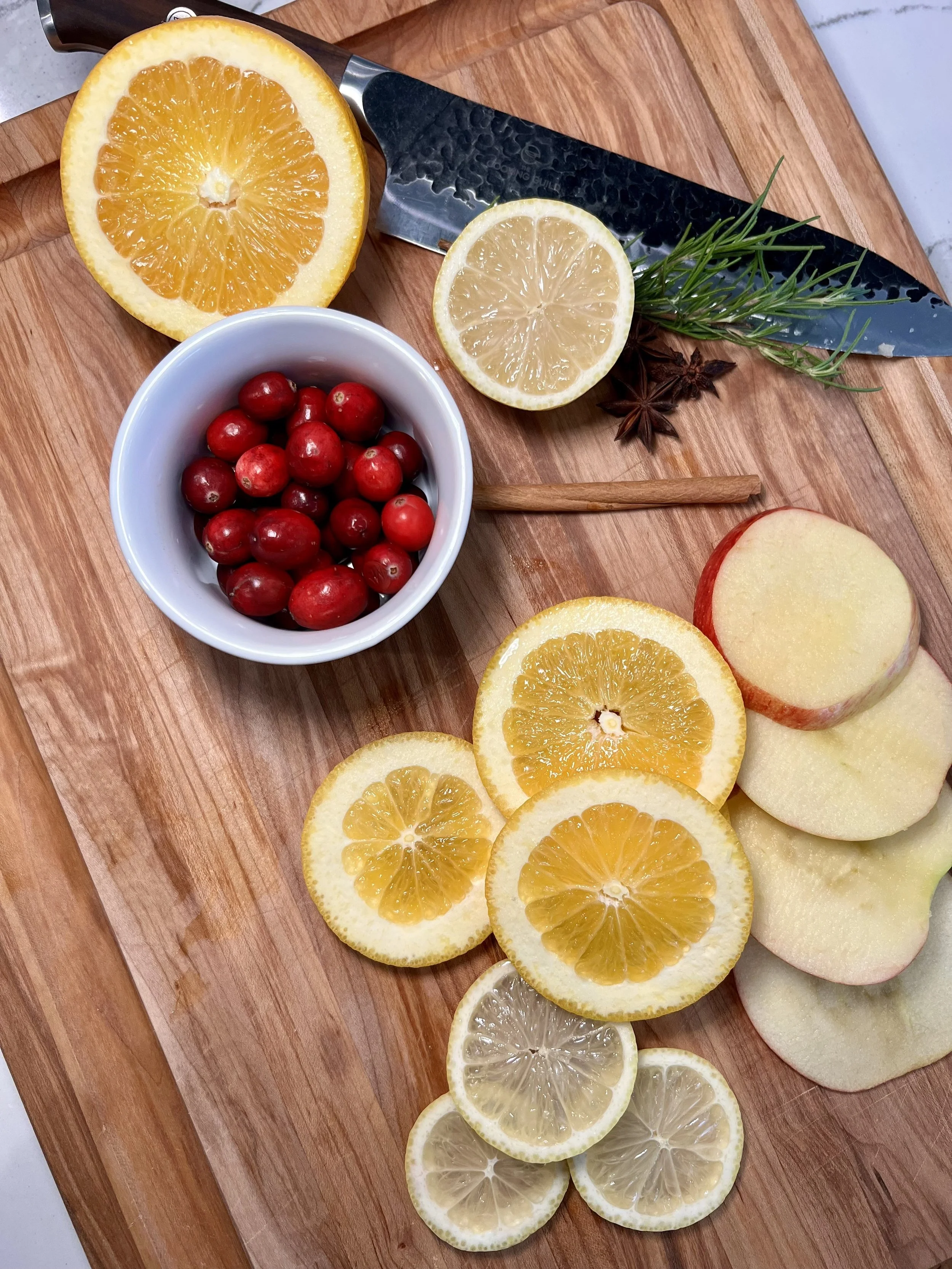 Wood Cutter board with sliced apples, lemon and oranges, small white bowl filled with cranberries along with a knife