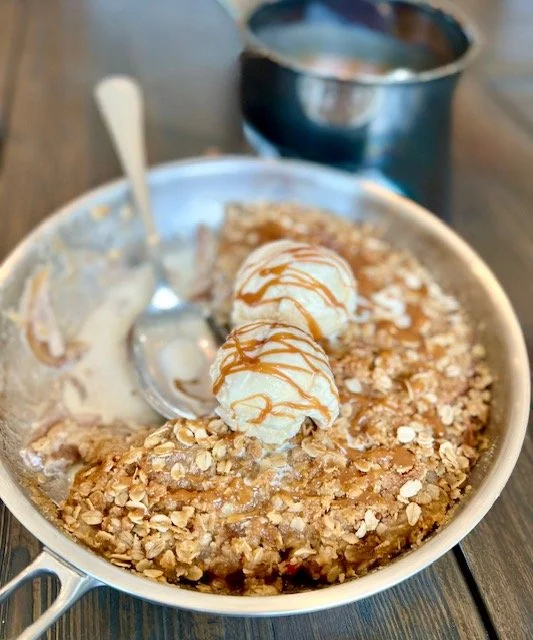 Skillet Pan filled with Apple Crisp, two scoops of vanilla ice cream in the middle with caramel drizzle. A spoon rests where some of the Apple Crisp as been served