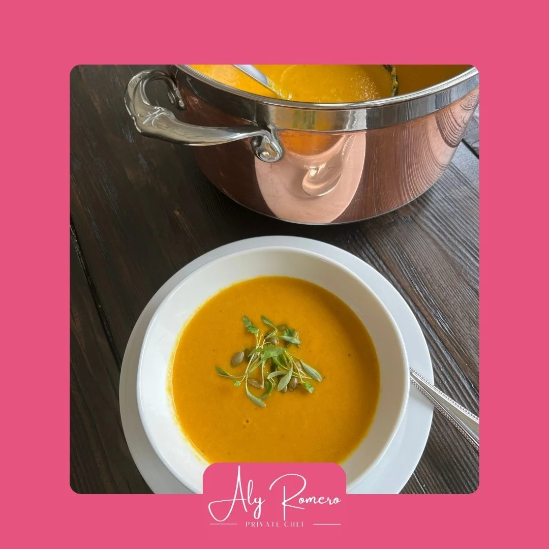A white bowl of orange-colored soup garnished with microgreens, on a dark wooden table, with a metallic soup pot in the background.