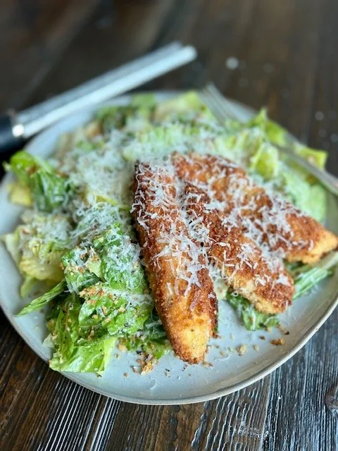 Salad with grilled chicken tenders, shredded cheese, and dressing on a white plate with white chopsticks on a wooden table.