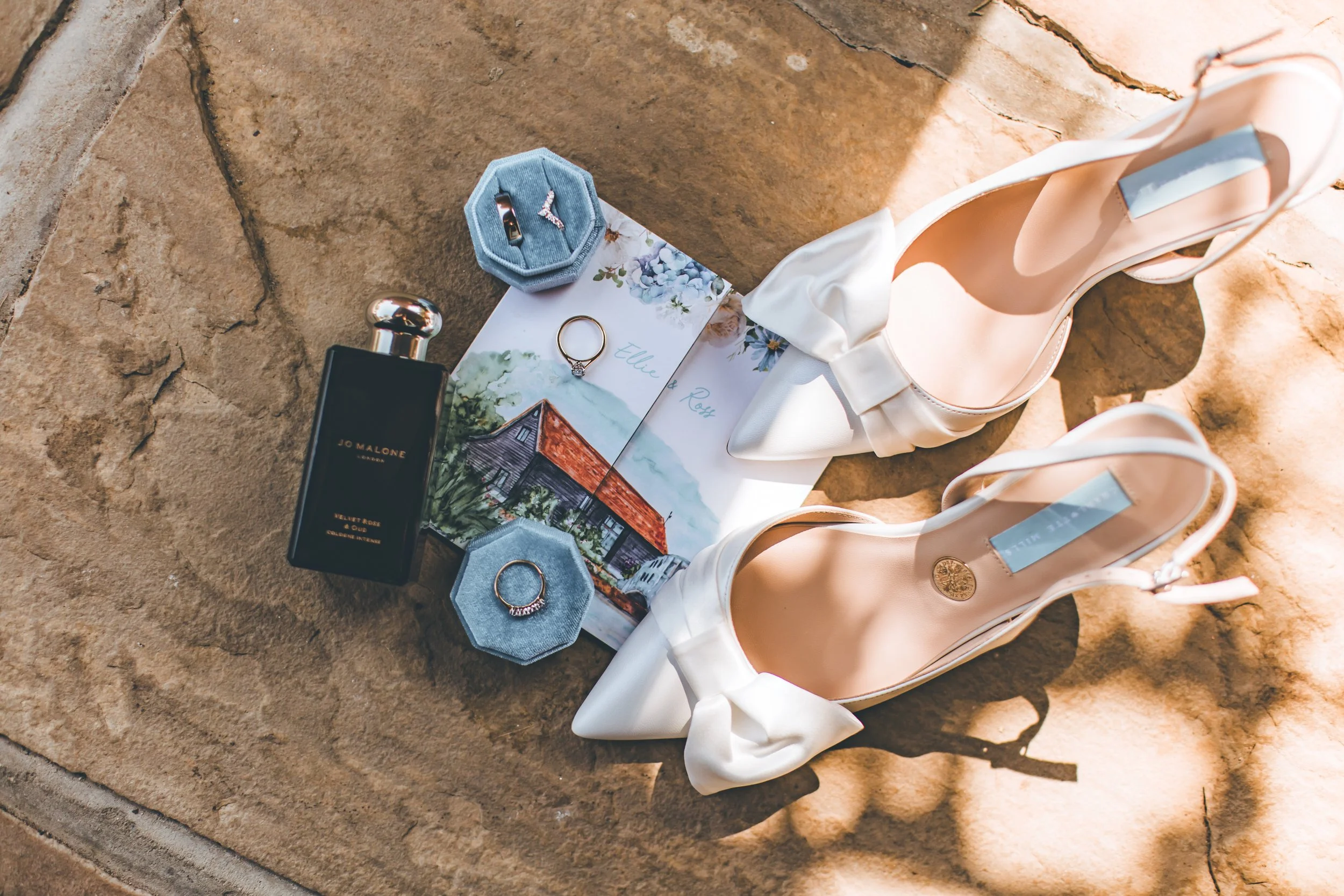 a wedding day flatlay including bridal shoes, perfume, rings and watercolour wedding invitation. Image by Shianne Mercer Photography
