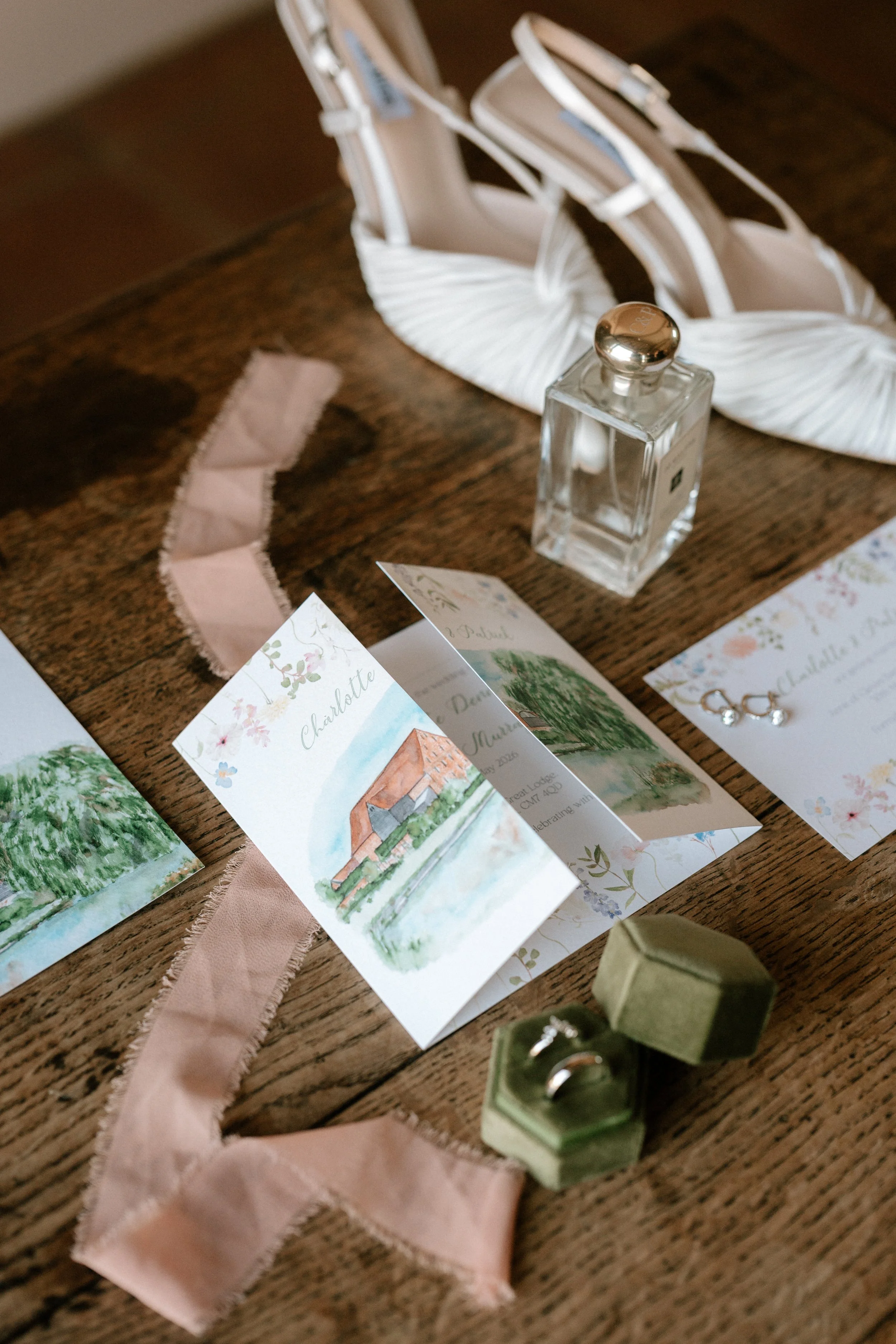 a wedding day flatlay including bridal shoes, rings & perfume and a watercolour gatefold invitation. Image by Laura Williams Photography.