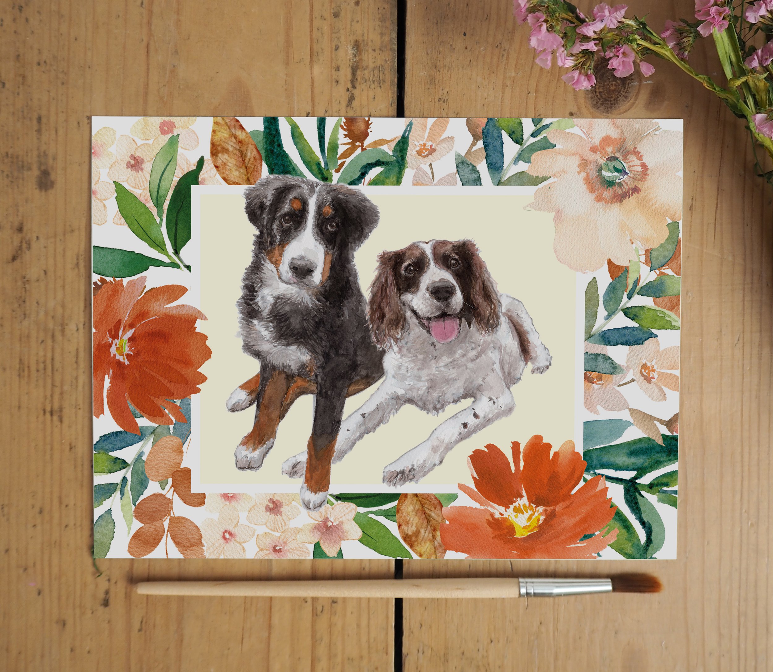 A watercolour print of 2 dogs on a yellow background with an autumnal floral border, on a wooden surface with paint brush and pink flowers