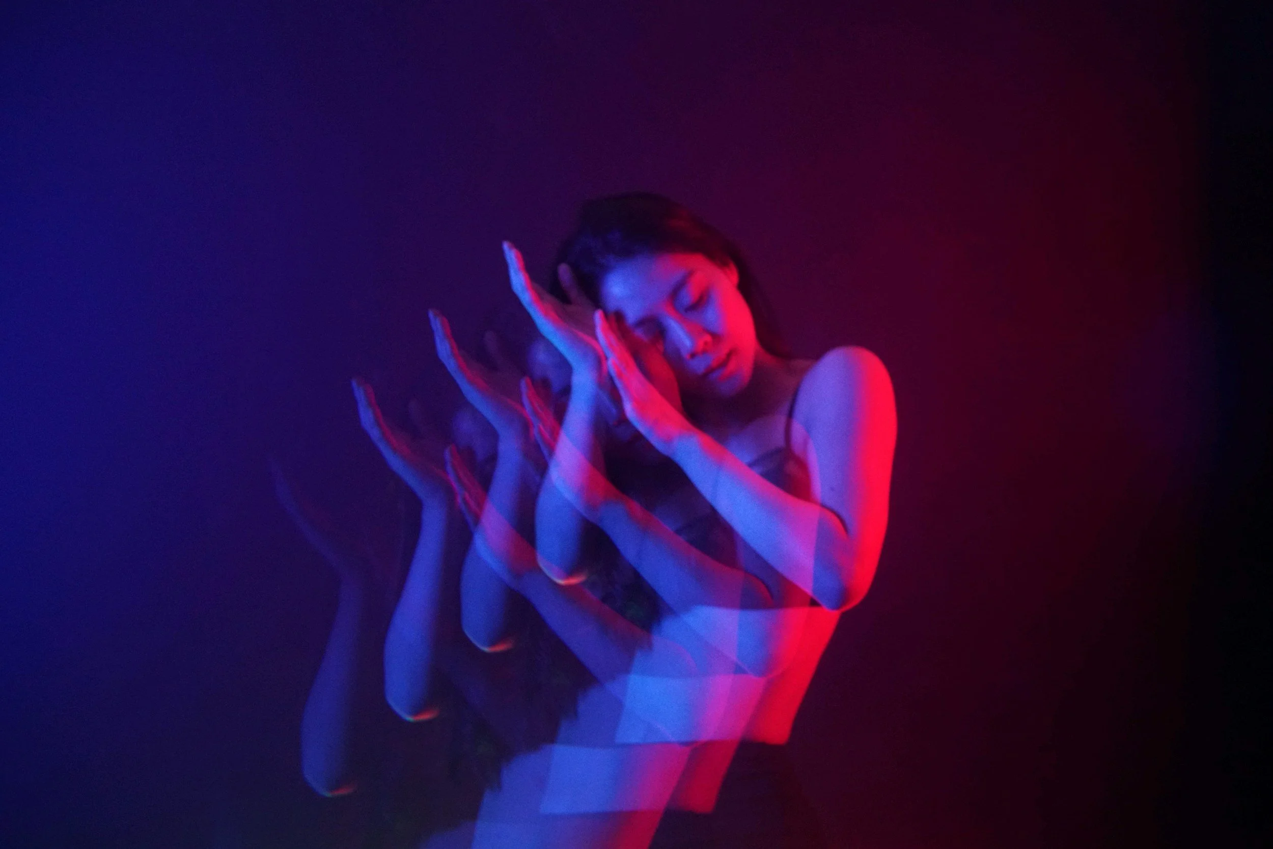 A woman with long dark hair posing against a dark background, with a multi-colored light effect creating a layered, moving appearance.