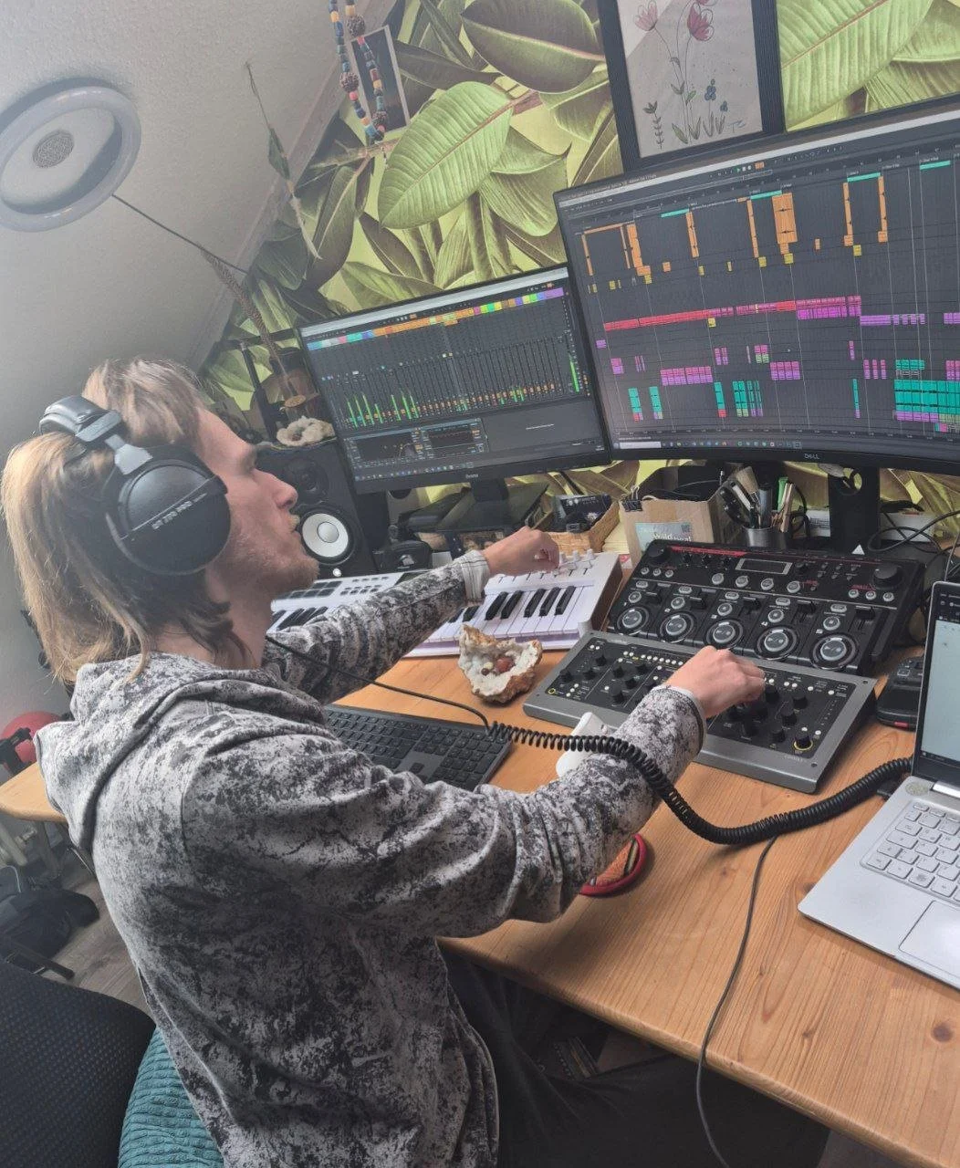 Ein junger Mann sitzt an einem Schreibtisch mit DJ-Equipment, mehreren Monitoren und Song-Software, trägt Kopfhörer und arbeitet an Musikproduktion, während er ein Stück Kuchen isst.