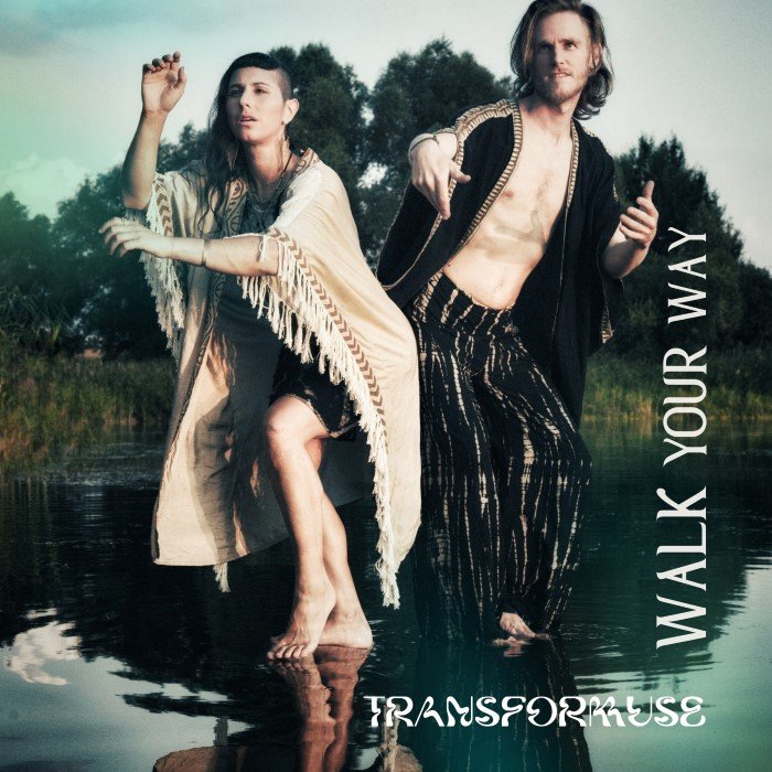 Ayam & ChrisTala standing in a body of water, with a natural landscape of trees and water behind them. Text Single's name overlay says 'WALK YOUR WAY'