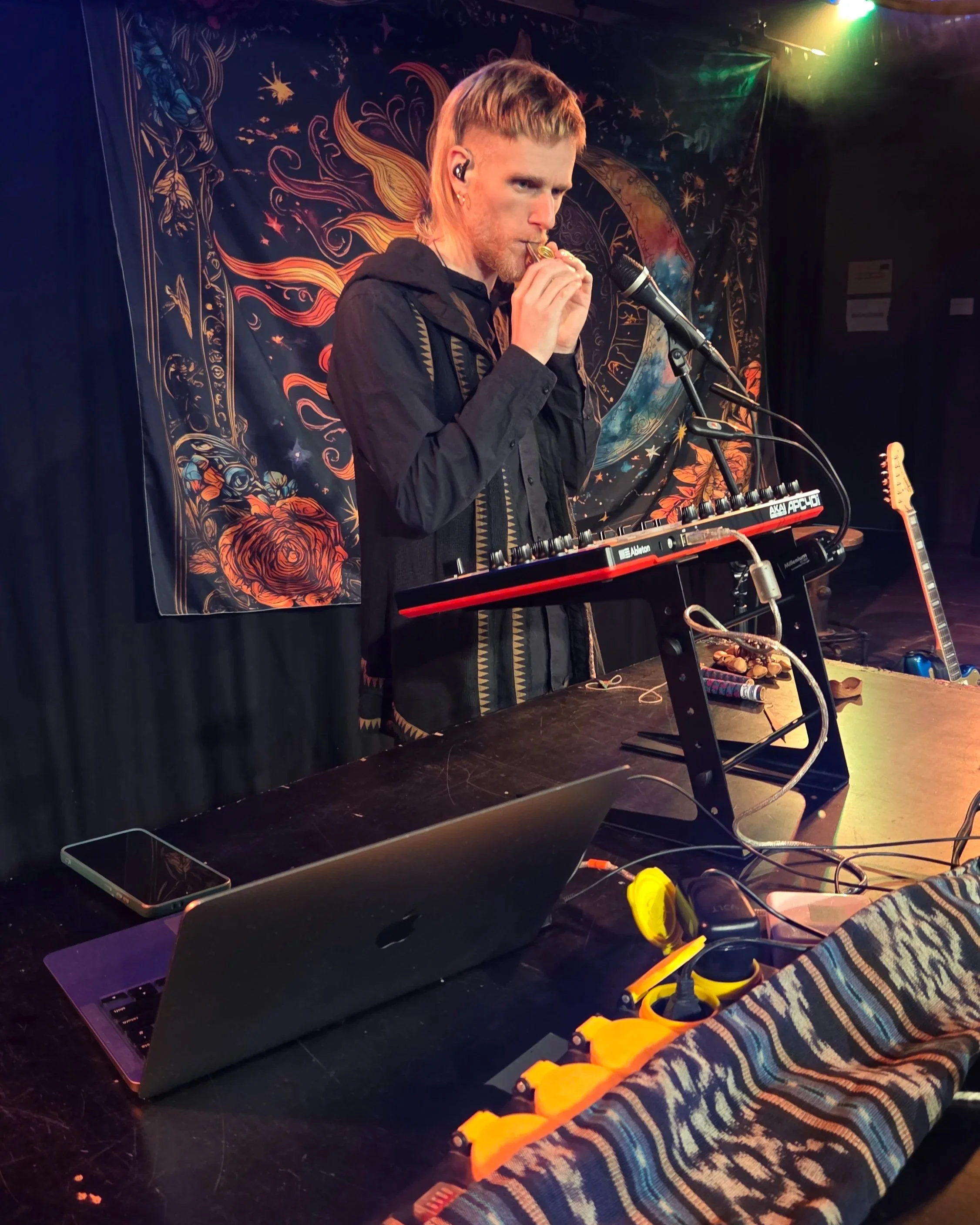 Ein junger Mann spielt ein melodisches Instrument auf einer Bühne mit buntem Sternenhimmel- und Feuerbogen-Hintergrund. Er trägt einen schwarzen Hoodie und hat kurze, blonde Haare. Es sind zudem ein Laptop, ein Aufnahmegerät und ein Gitarre im Bild.