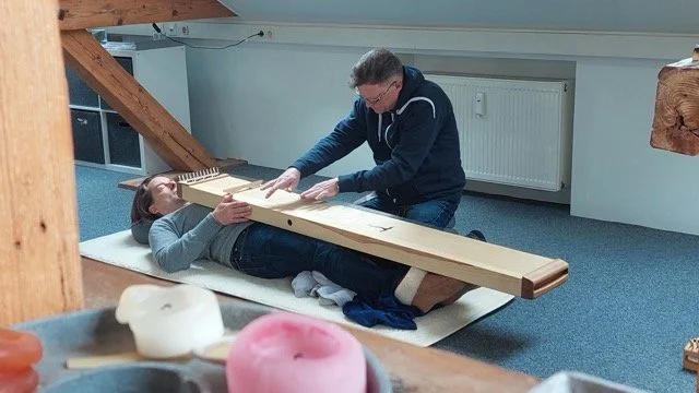 Ein Mann legt eine Frau auf den Rücken auf eine Matte, während er eine große Holzbank auf den Bauch der Frau legt.
