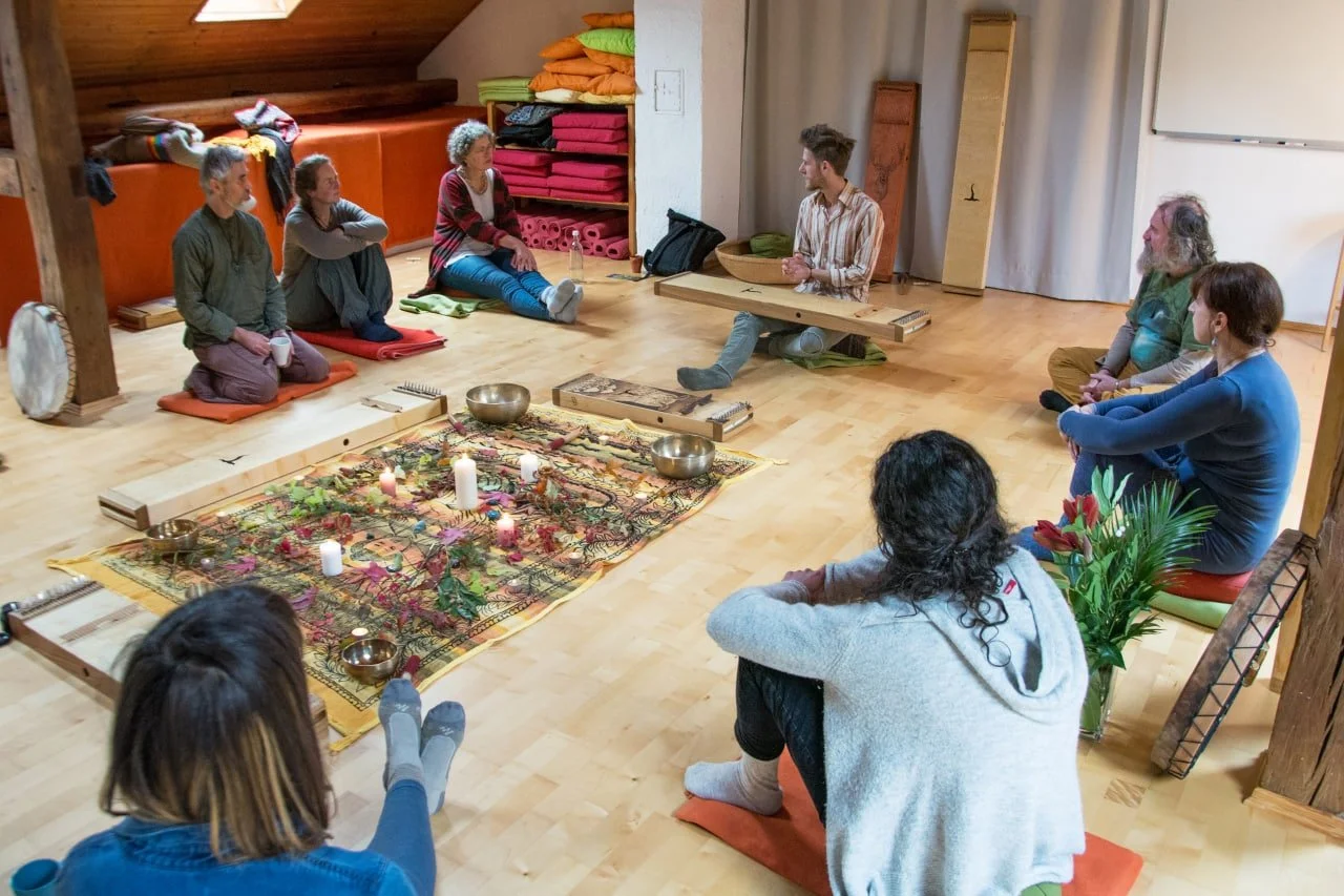 Eine Gruppe von Menschen sitzt im Kreis auf Kissen auf dem Holzboden, um eine Meditation oder spirituelle Zeremonie. Im Mittelpunkt befindet sich eine bunte Decke mit Kerzen und Pflanzen.