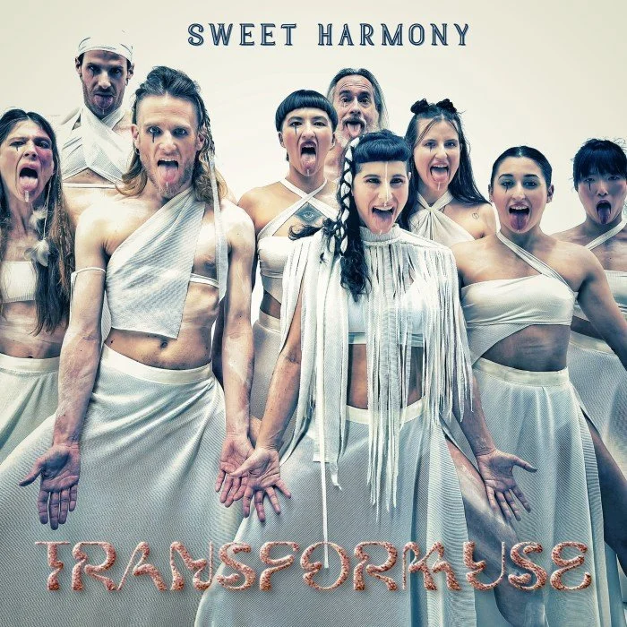 Group of people dressed in white, theatrical costumes, with their tongues out, standing in front of a sign that says 'SWEET HARMONY' and 'TRANSPORRUSE' at the bottom.
