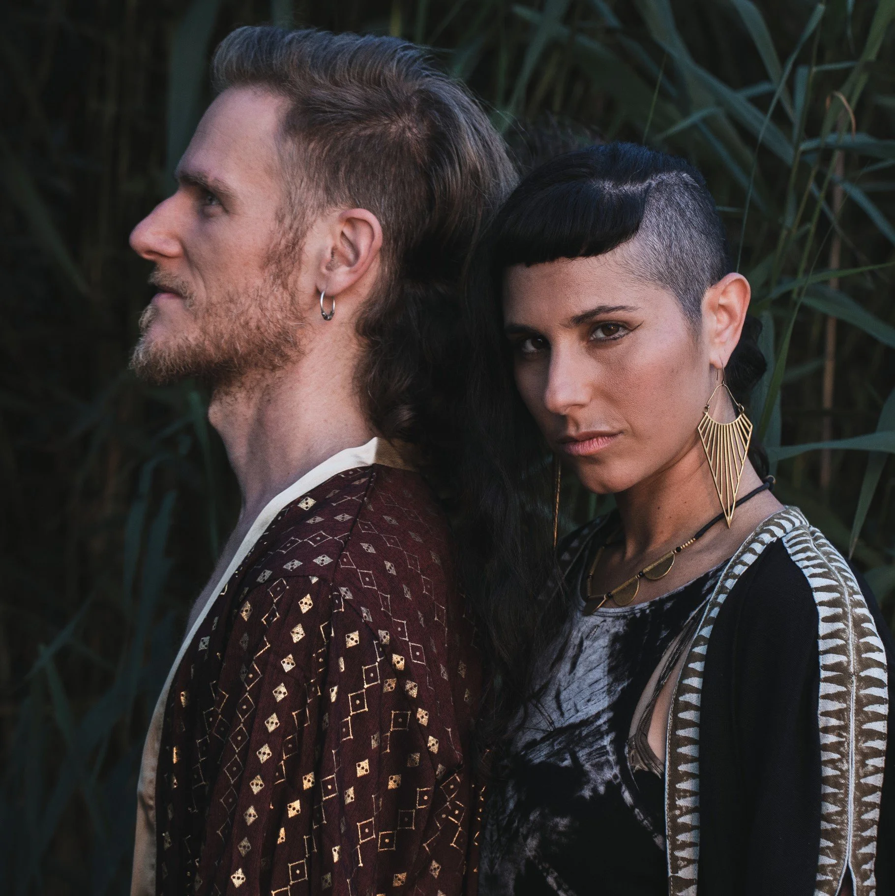 ChrisTala with a beard and earring faces left with eyes closed, and Ayam with short dark hair and large earrings faces right, standing close with their backs touching in a natural outdoor setting with tall green plants.