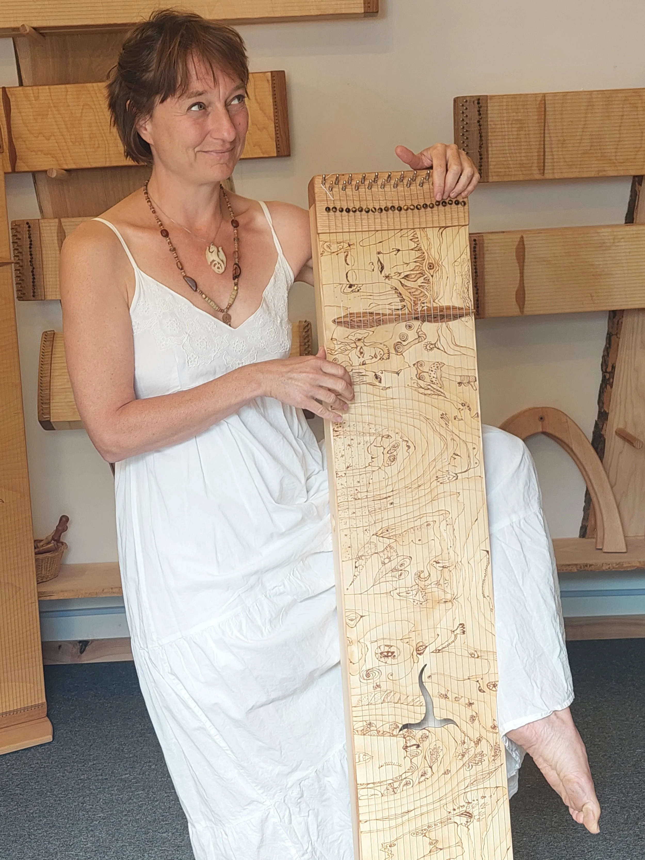 Eine Frau trägt ein weißes Kleid und sitzt auf einem Stuhl. Sie hält ein Holzbrett mit geschnitzten Tierbildern und Linien, die Tierwanderungen darstellen. Im Hintergrund sind Holzregale an der Wand.