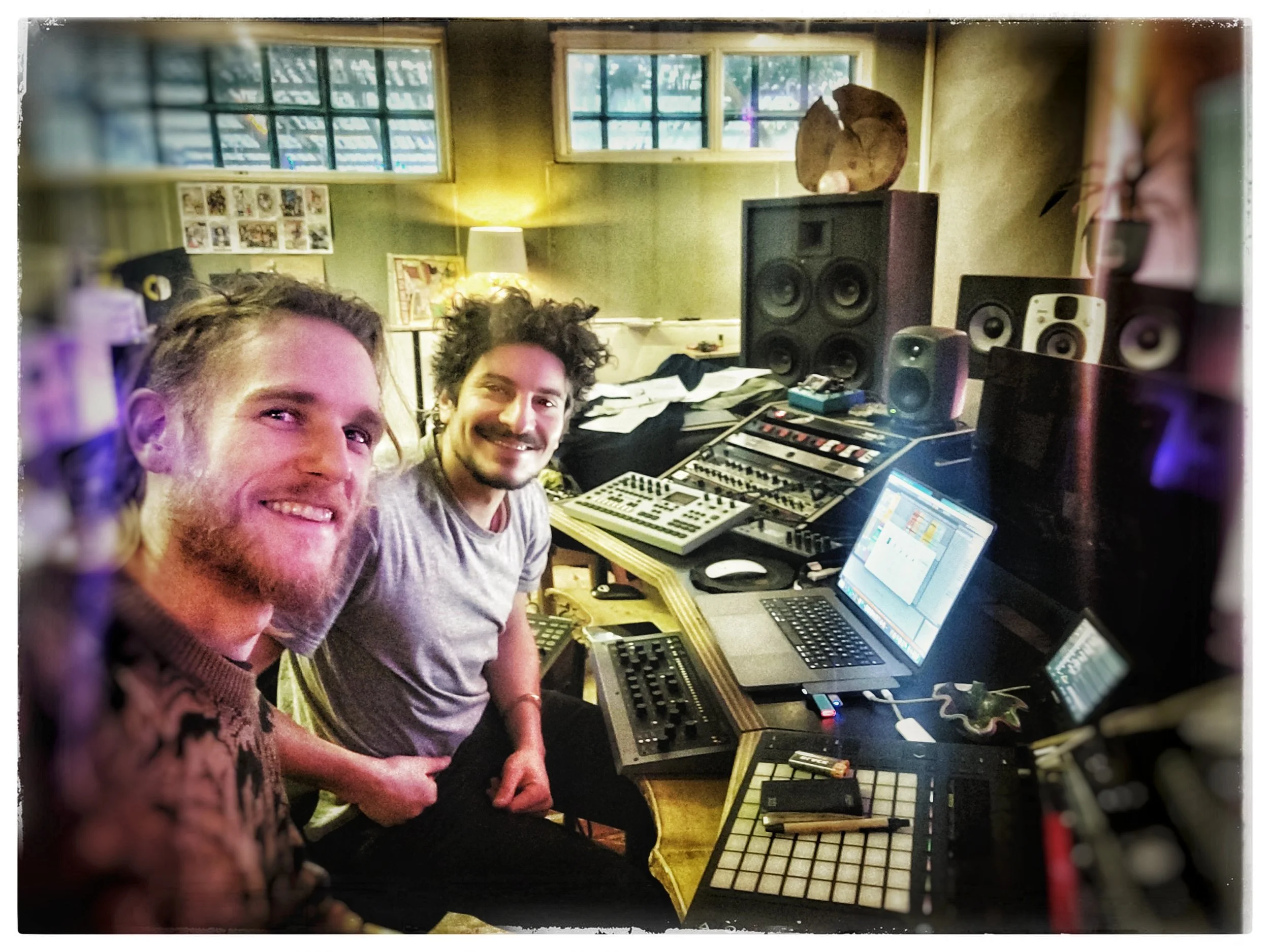 Zwei Männer sitzen in einem Musikstudio mit elektronischen Geräten, Monitoren und Lautsprechern und lächeln in die Kamera.