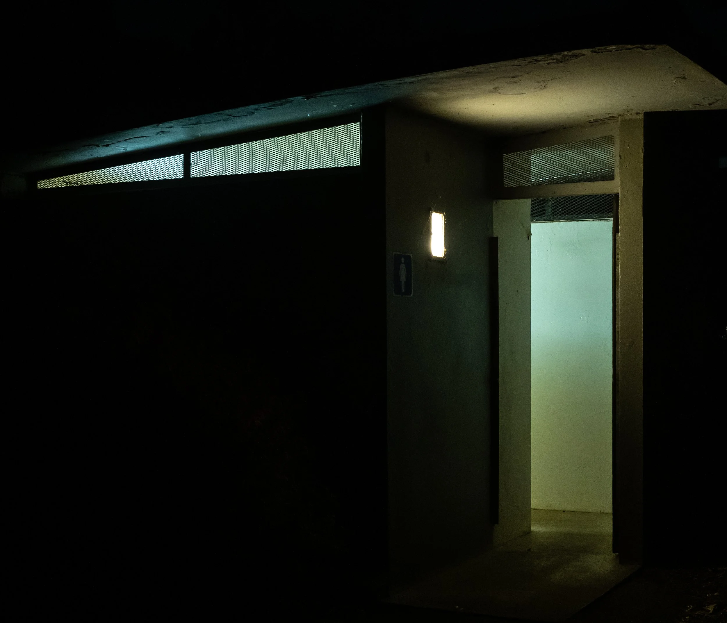 Dark building entrance with a lit doorway and small window panels near the ceiling.
