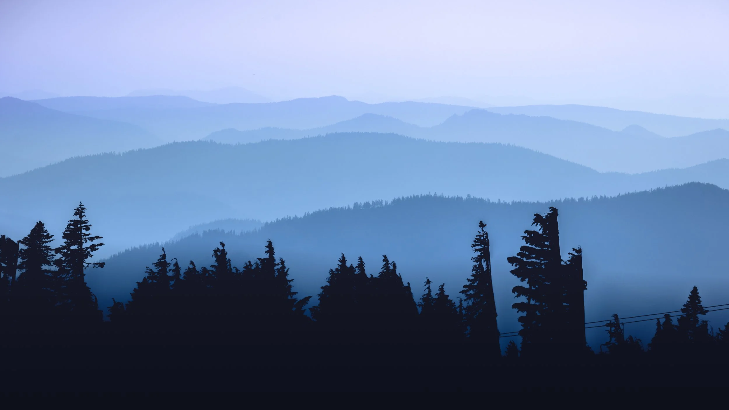 Silhouetted trees in front of layered blue mountain ranges with a light sky.