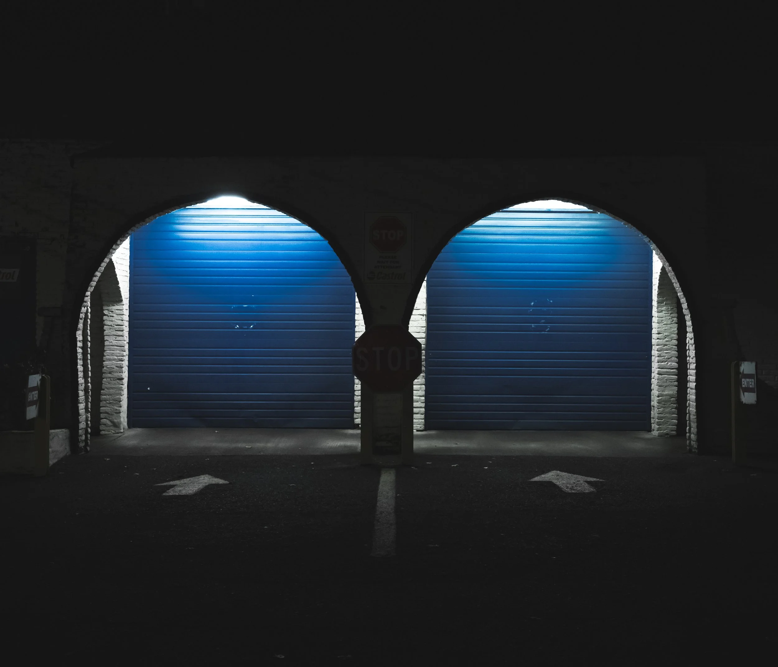 Night view of two closed blue garage doors with white arrows on black pavement pointing right, under a dark archway, with stop signs and exit signs on the sides.