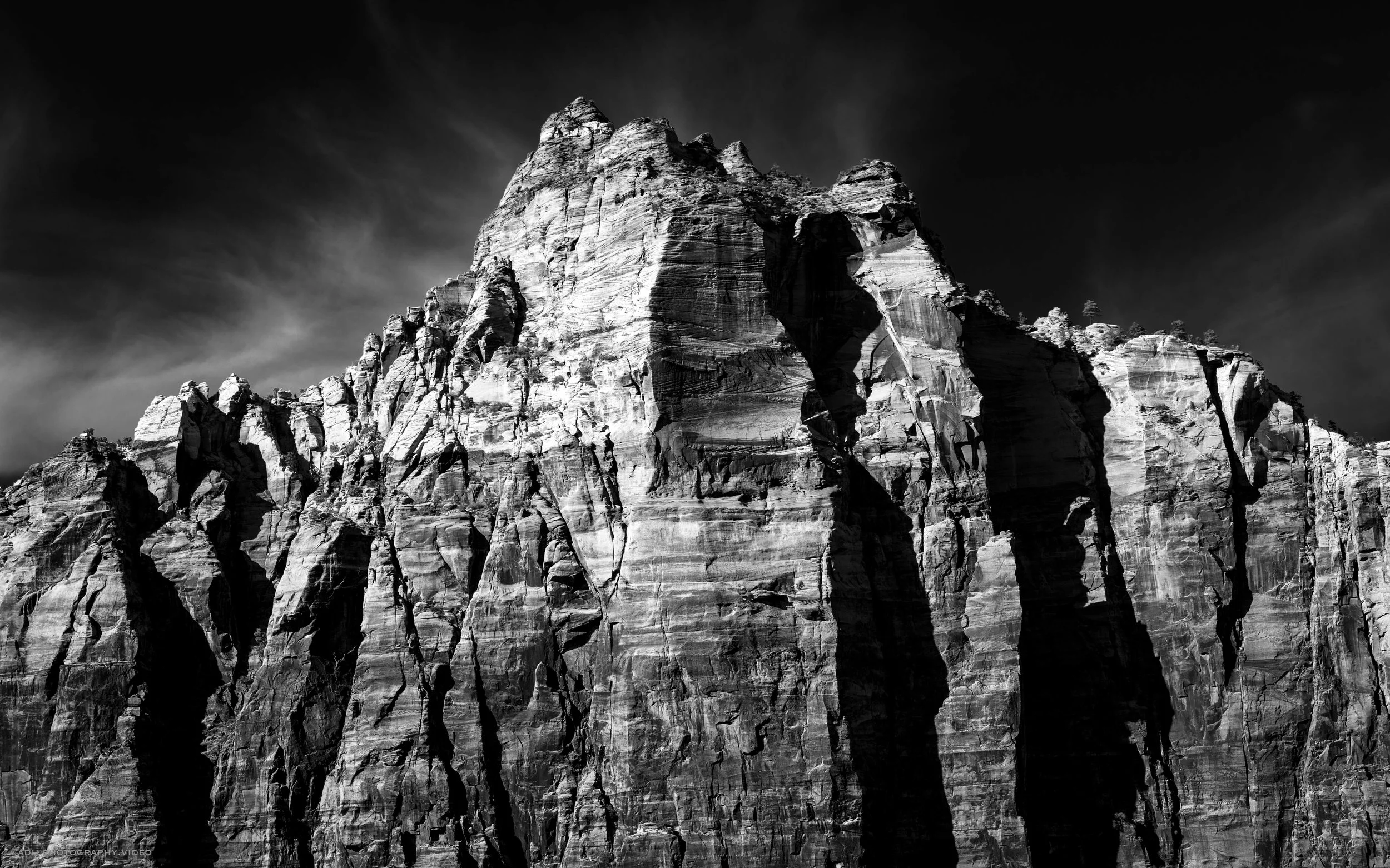 Black and white photograph of a tall, rugged mountain with steep cliffs and jagged rocks under a dark sky.