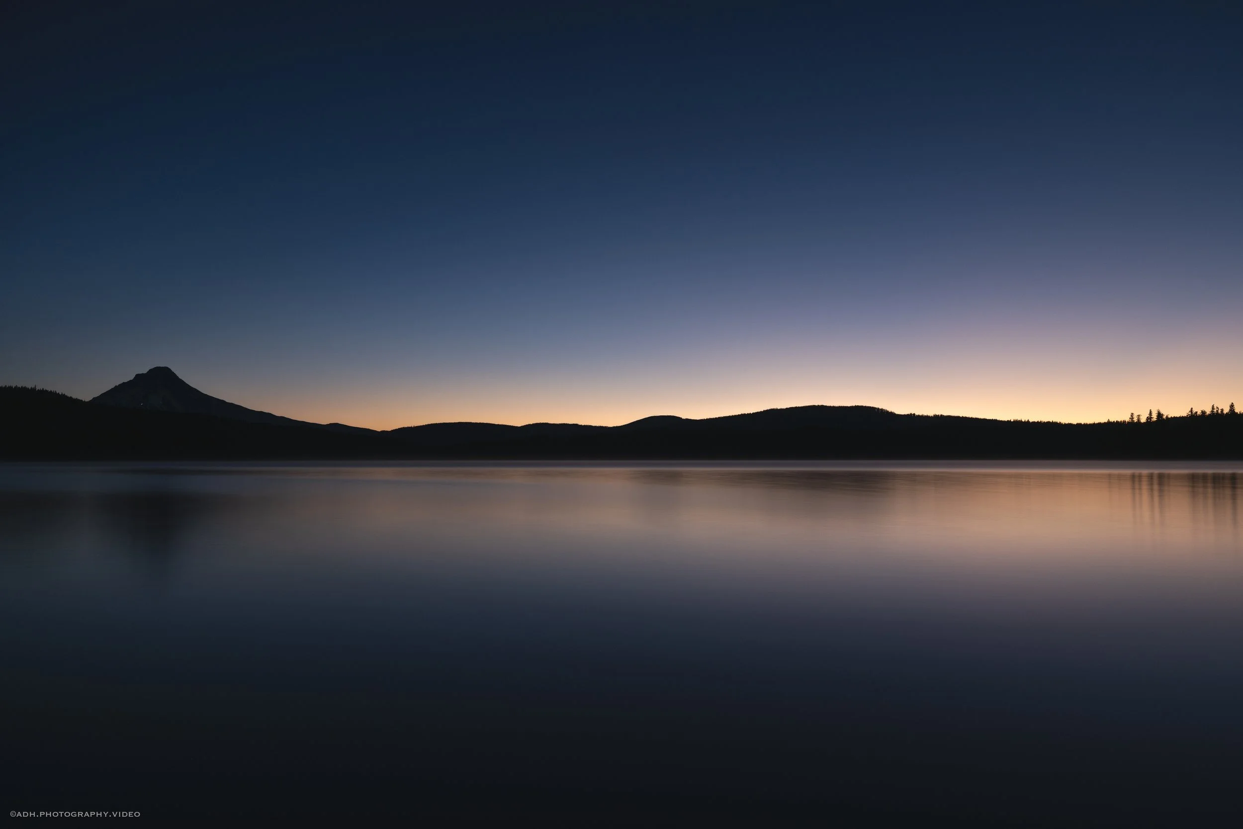 Serene lake at dusk with calm water reflecting dark silhouettes of distant hills and a mountain with a pointed summit on the left, under a fading gradient sky from pale yellow to deep blue.