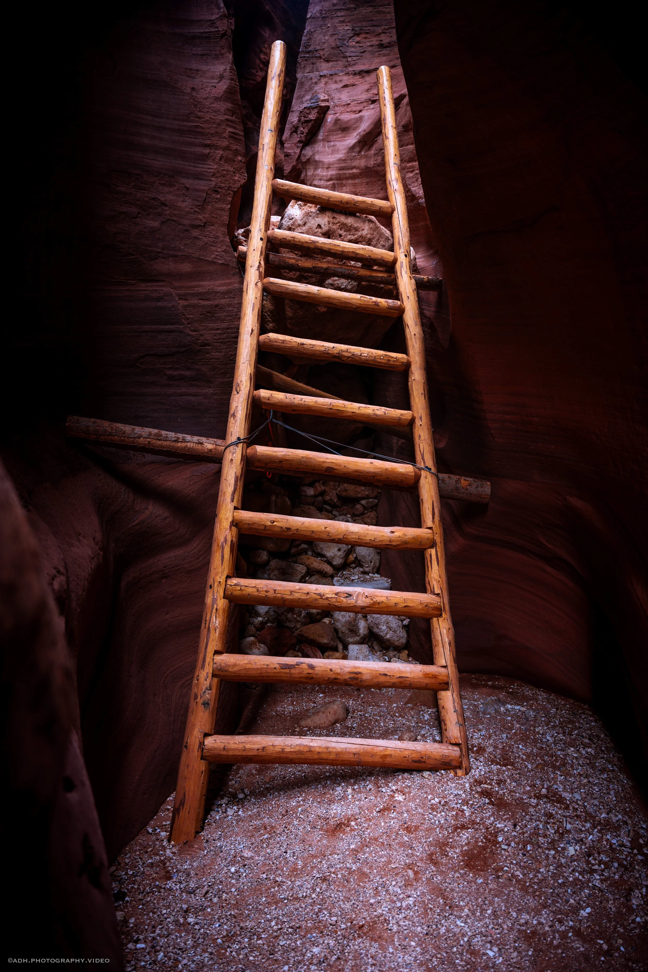 A wooden ladder leaning against the sandstone walls of a narrow canyon with a rocky floor.