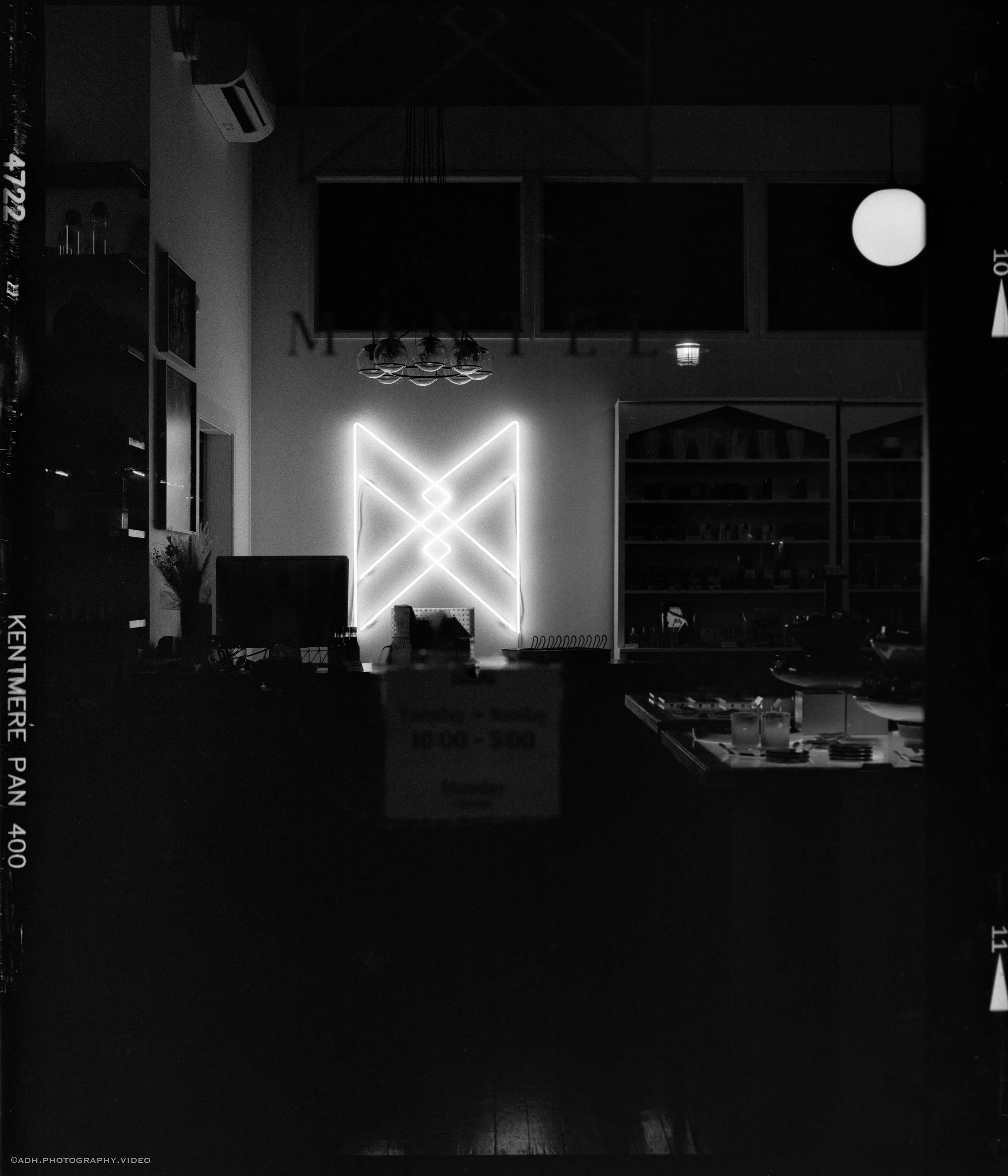 Interior of a dimly lit room with a neon geometric wall art centerpiece, shelves with books, a desk with a computer, and hanging light fixtures.