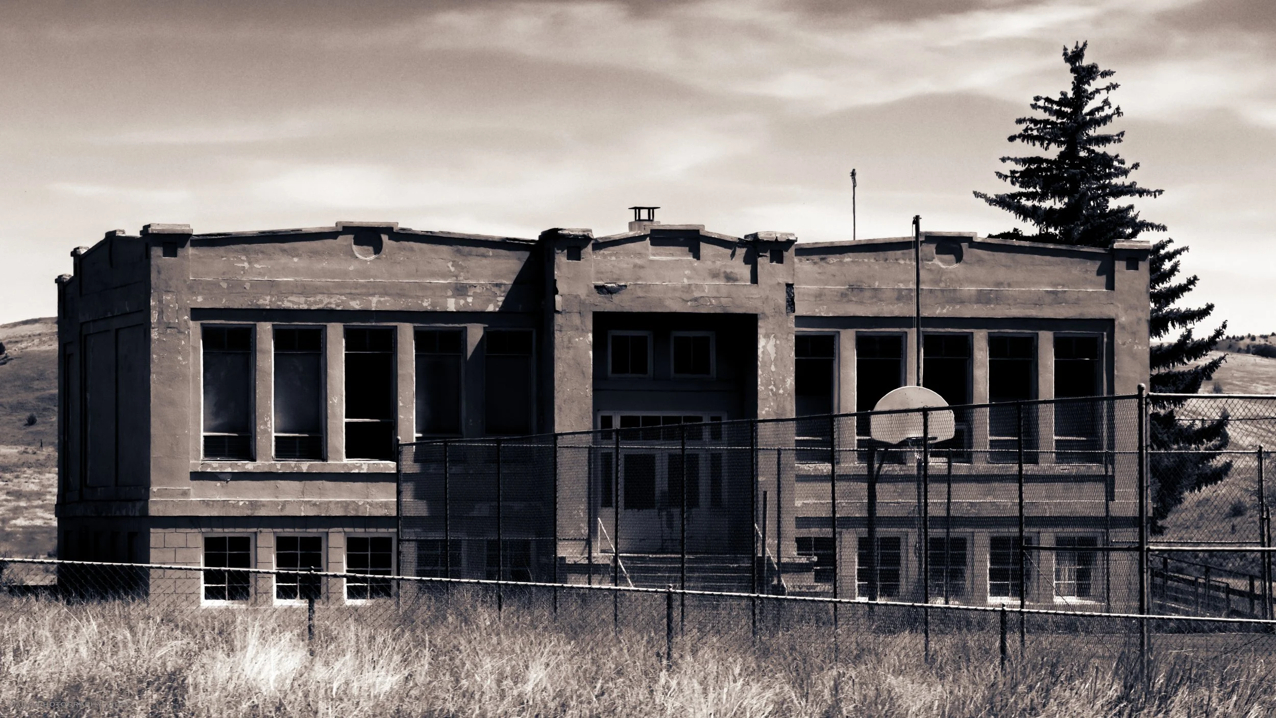 An abandoned, weathered basketball court area with a backdrop of a large, dilapidated building with broken windows, surrounded by a chain-link fence, and a solitary tree in the background.