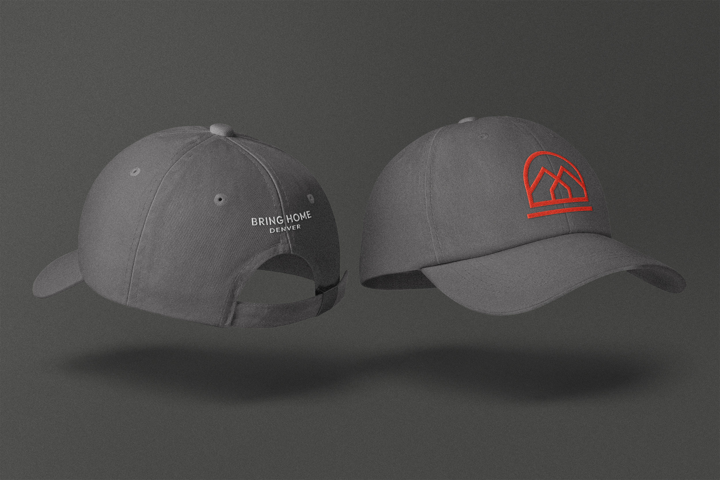 Two gray baseball caps suspended in midair against a dark gray background. The cap on the left features white embroidered text that reads 'BRING HOME DENVER' on the back, with a small adjustable strap. The cap on the right has a red geometric logo on the front.
