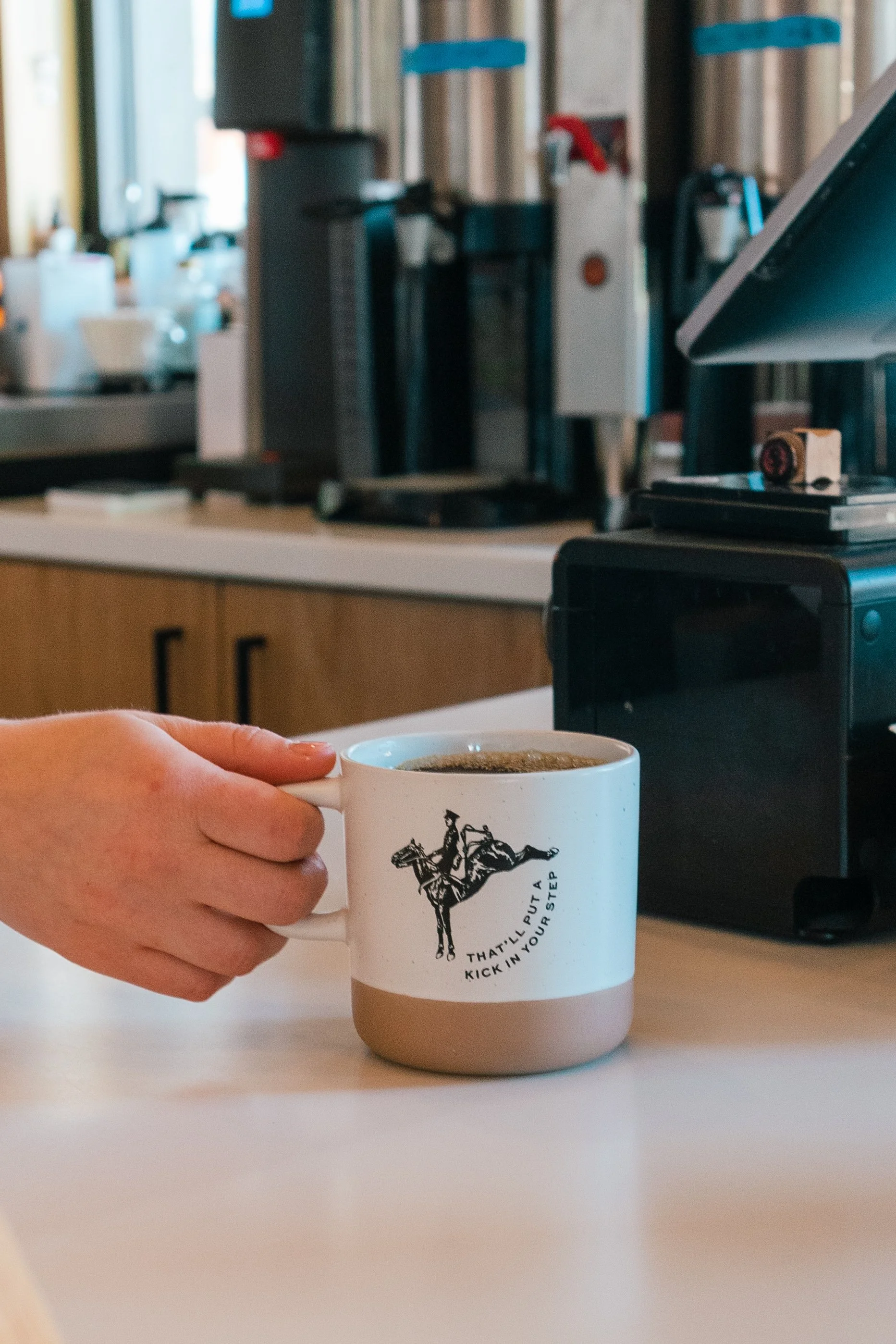 A person holding a white coffee mug with a rider on a horse and the text 'That'll put a kick in your step' on a kitchen counter.