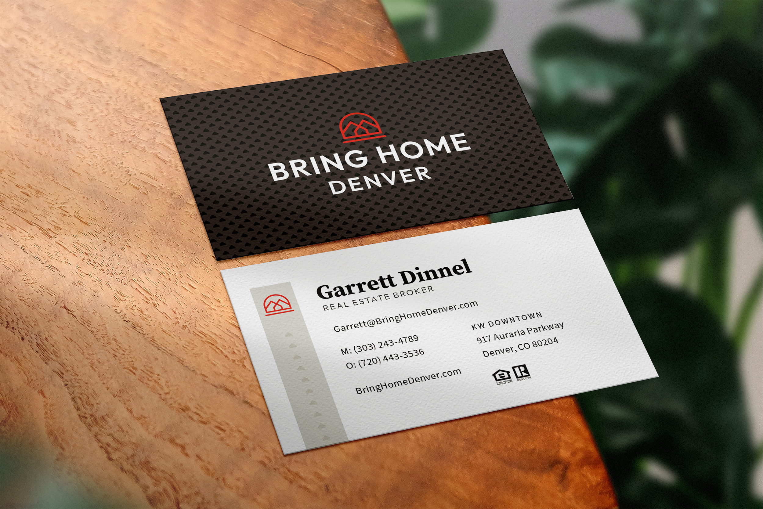 Business card for Garrett Dinnel, a real estate broker, with contact information, branding logo, and slogan 'Bring Home Denver'. The card is on a wooden surface with blurred green foliage in the background.