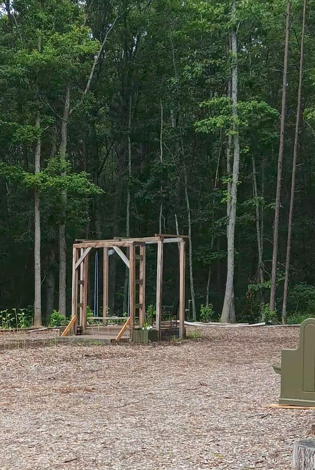 A wooden play structure with swings in a wooded outdoor area with trees and mulch covered ground.