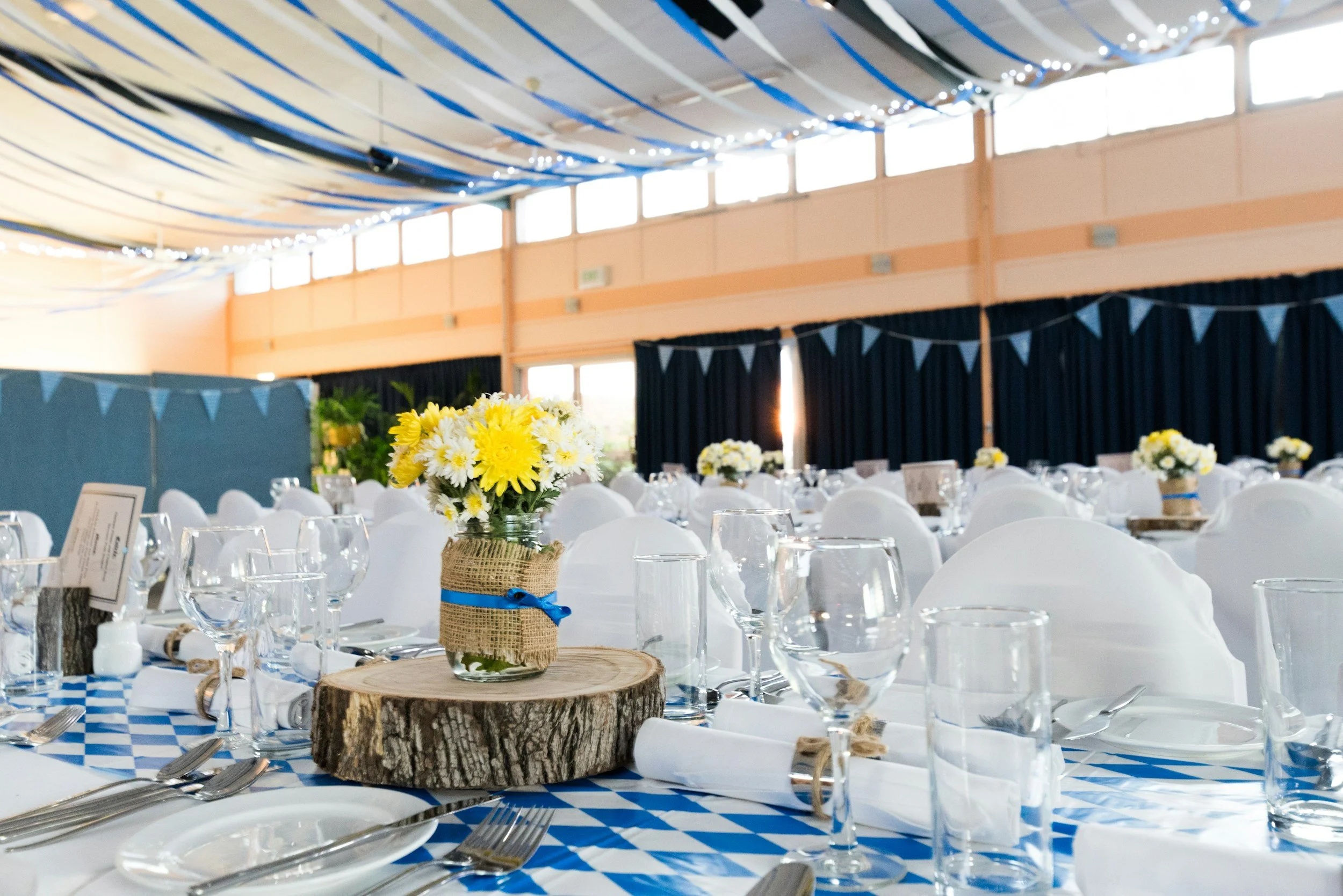 Decorated event hall with tables set for a formal occasion, featuring floral centerpieces with yellow and white flowers, blue and white tablecloths, white chair covers, and hanging blue streamers and bunting.