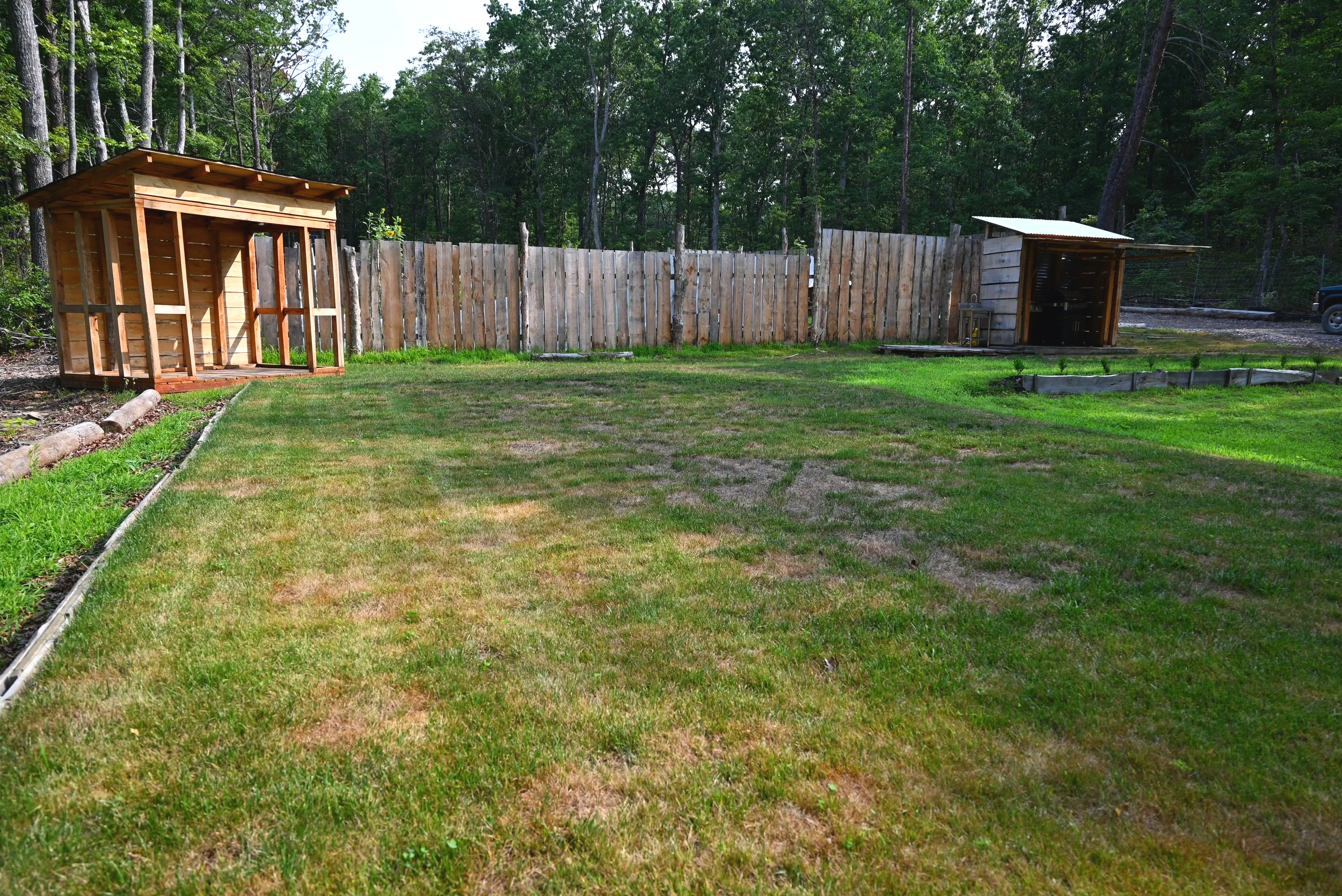 Backyard with green grass, wooden fence, and two small wooden structures with open ends, surrounded by trees.