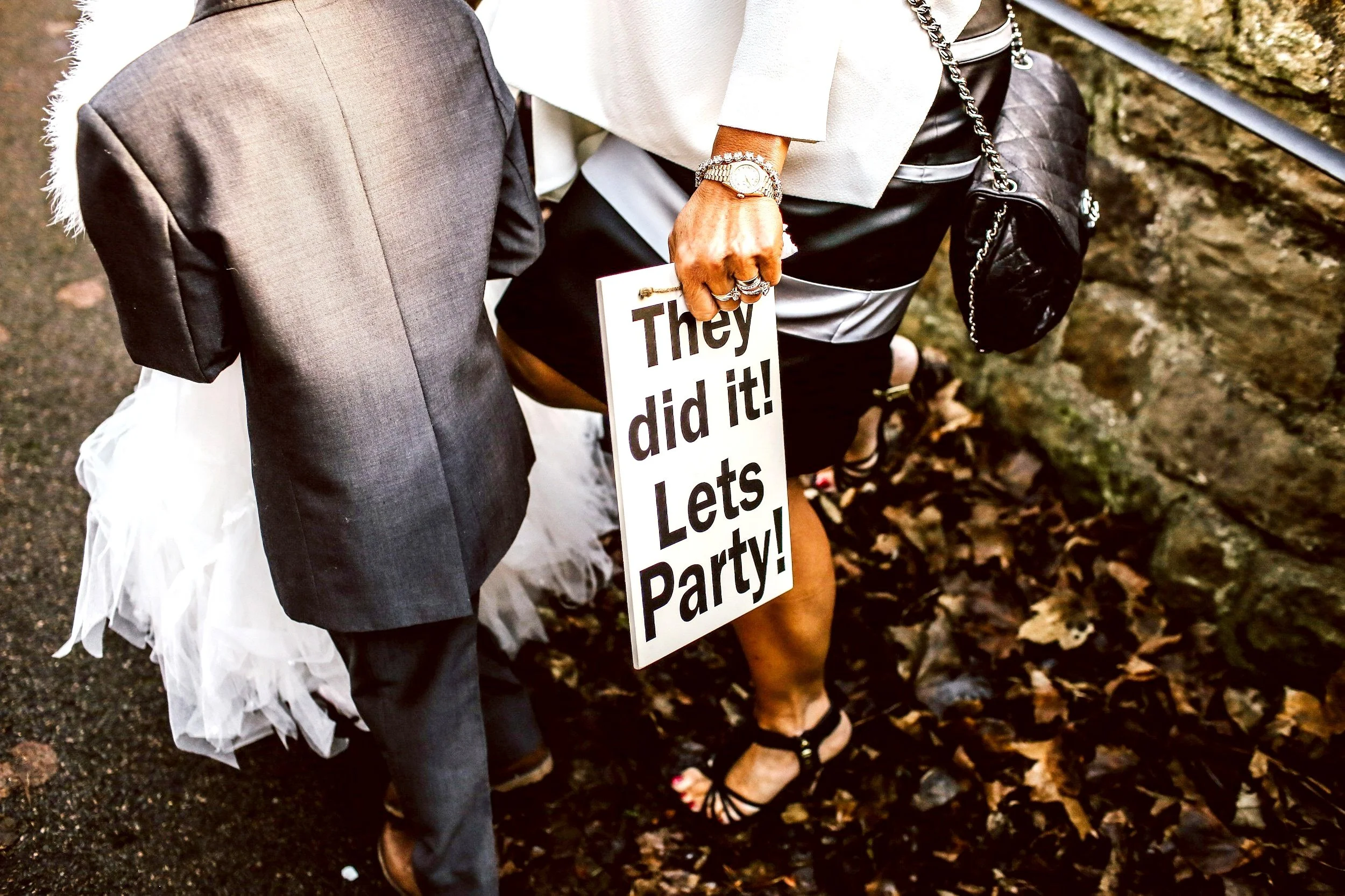 Person holding a "They did it! Lets Party!" sign and a child in formal attire standing on leaves.