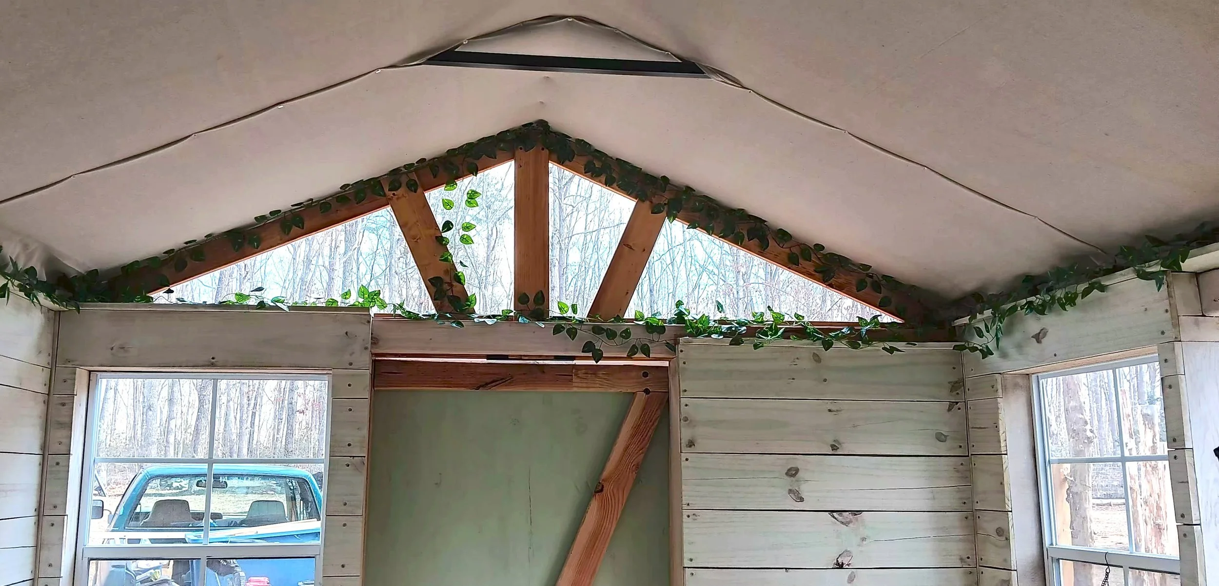 Interior of a wooden A-frame structure with unfinished walls and ceiling, decorated with artificial green vines. Two windows are visible, one on each side, showing a snowy outdoor scene with trees. A vehicle is seen through the left window.