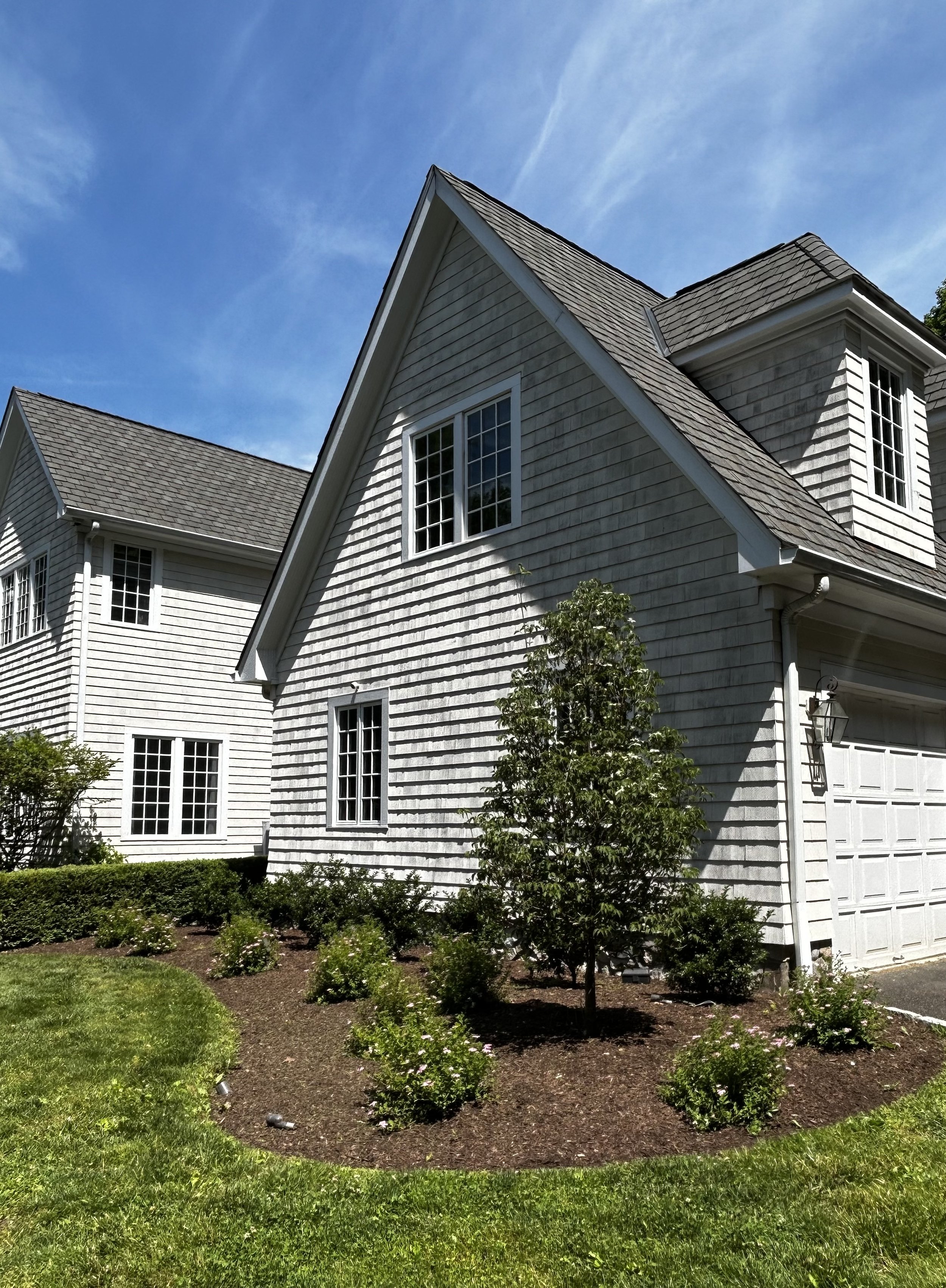 A white two-story house with gray shingles, multiple windows, a garage door, and a front yard with green grass, shrubs, and small plants under a bright blue sky with wispy clouds.