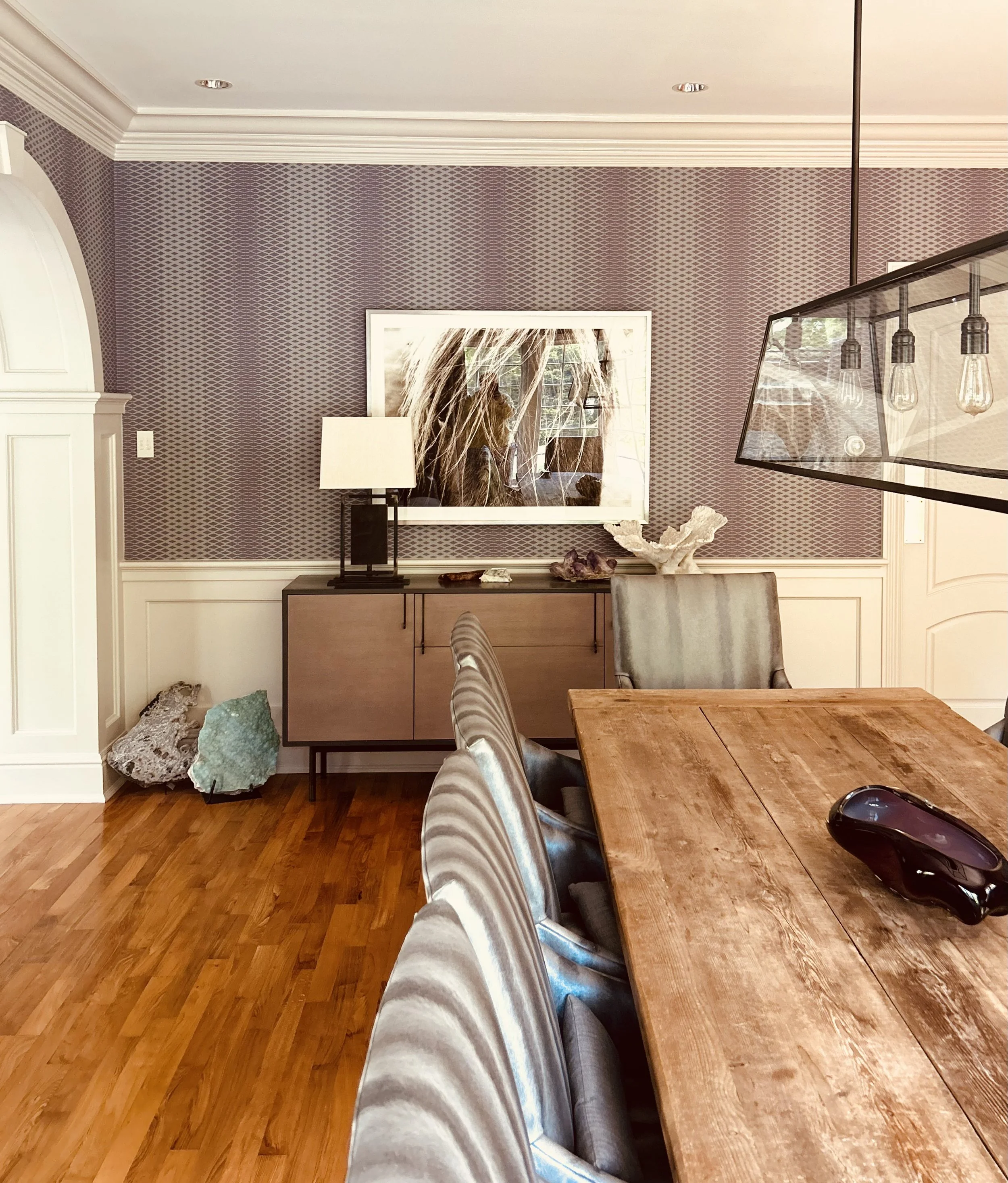 Person sitting at a wooden dining table with metallic legs, holding a smartphone in one hand, with a black vase or bottle on the table, in a room with patterned purple wallpaper, a large mirror reflecting a window, a beige and black table lamp, and a