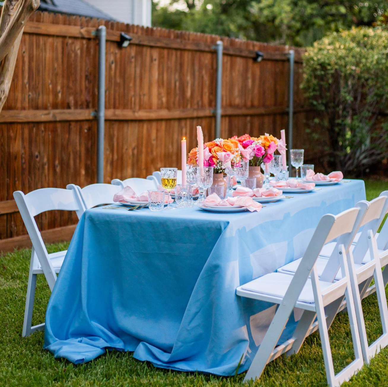 Outdoor dining table decorated for a celebration with pink floral centerpieces, pink napkins, candles, and glassware, set on a blue tablecloth in a backyard with a wooden fence and greenery.