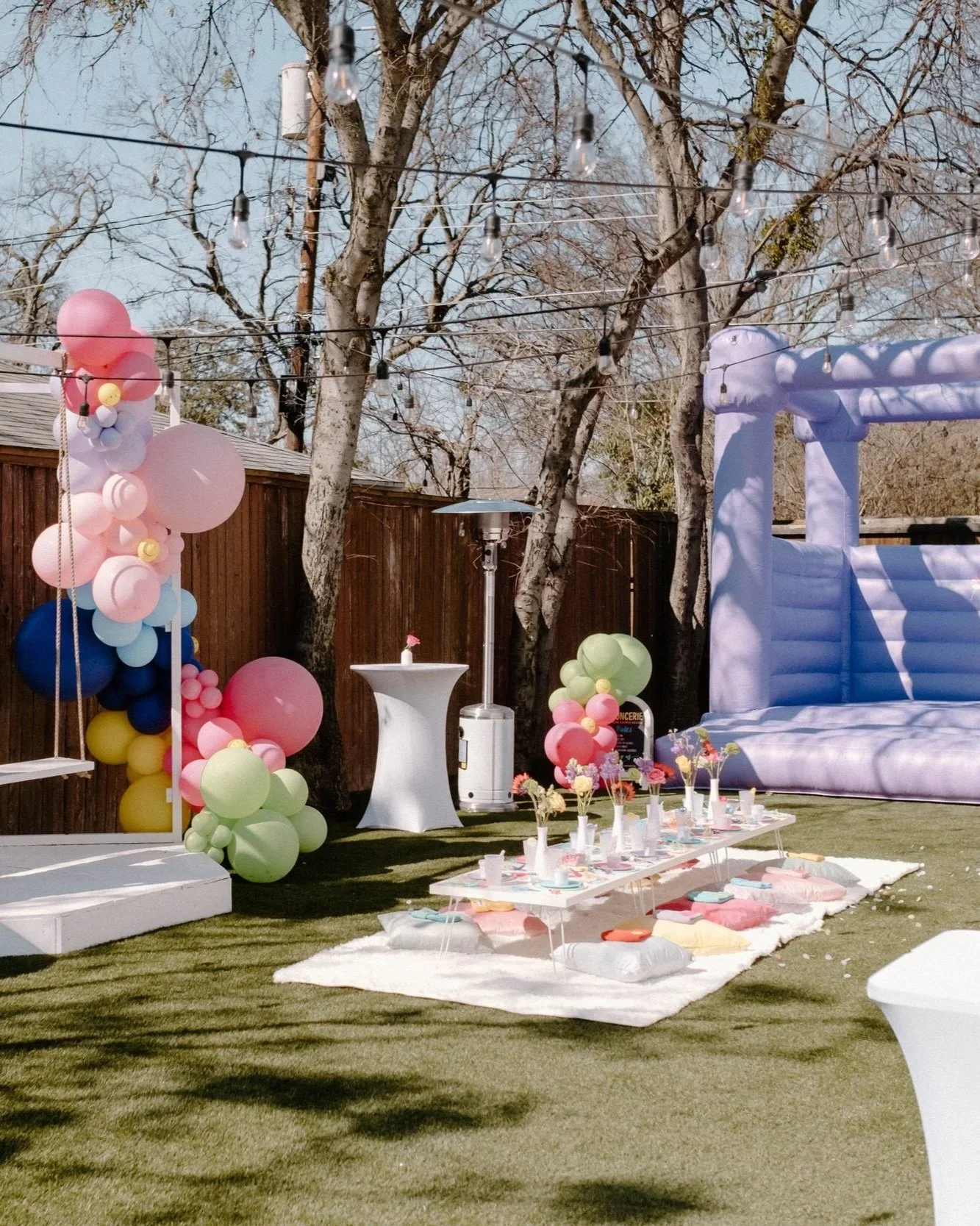 Decorated outdoor space with pink, lavender, yellow, blue, and green balloons, a white table set with plates and flowers, a purple inflatable structure, string lights, and trees in the background.
