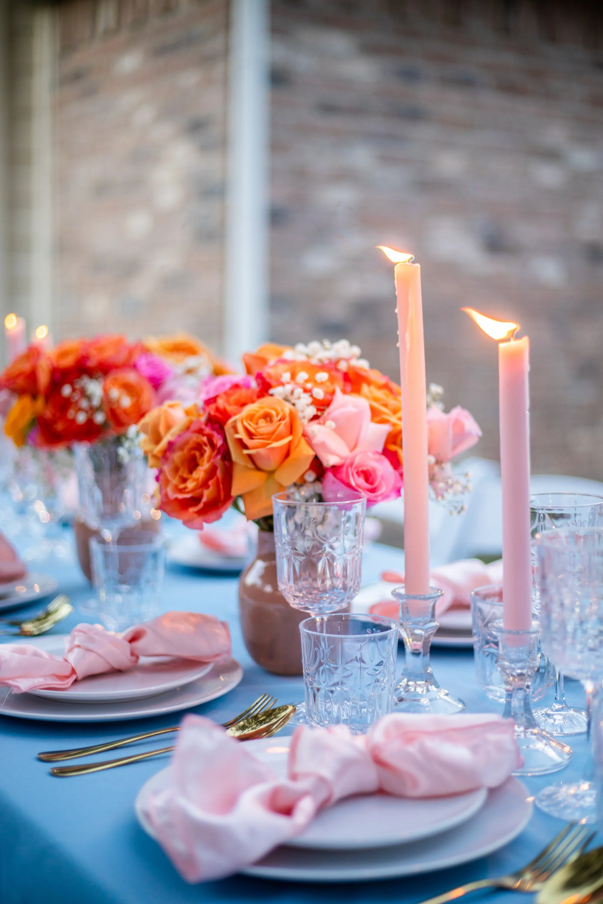 Elegant table setting with pink candles, peach and pink roses, and gold cutlery on a blue tablecloth.