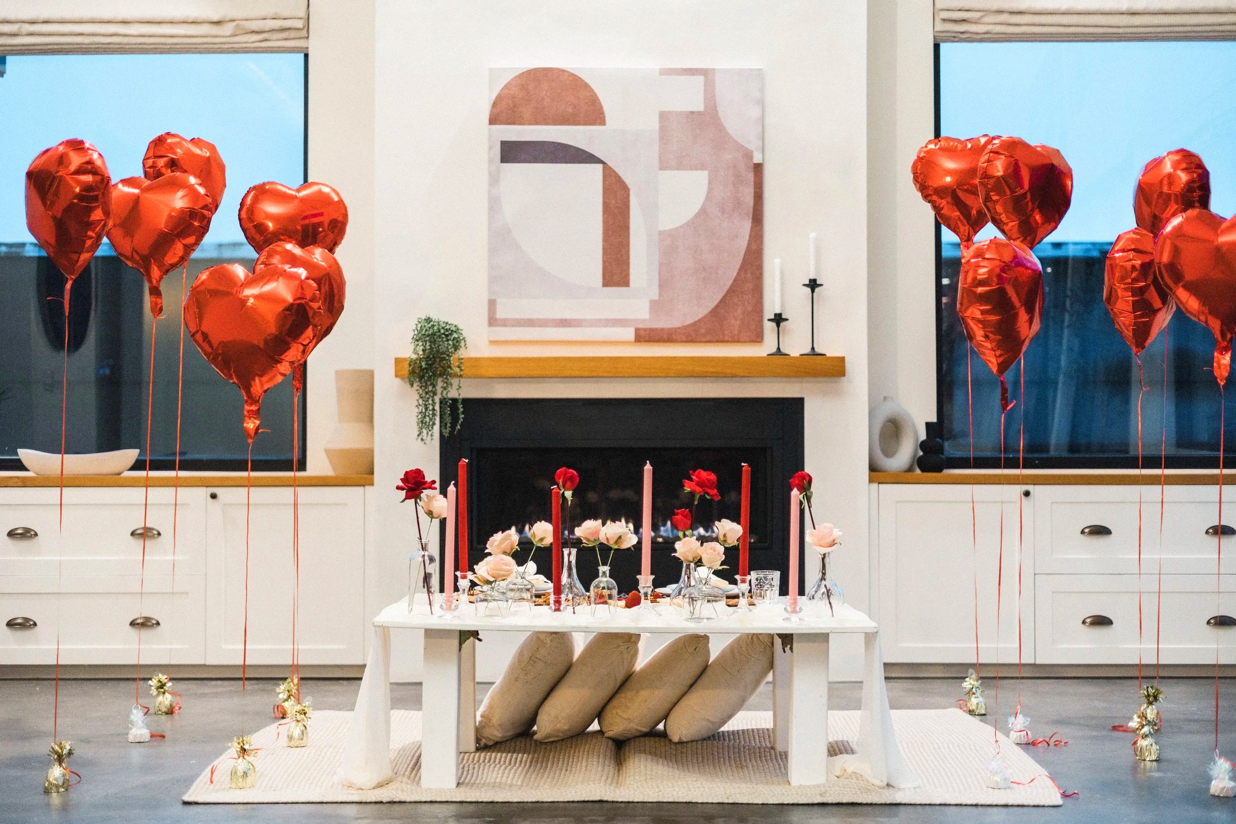Valentine's Day decorated living room with heart-shaped red balloons, a white coffee table with pink and red roses, candles, and birthday favors, in front of a fireplace and abstract artwork.
