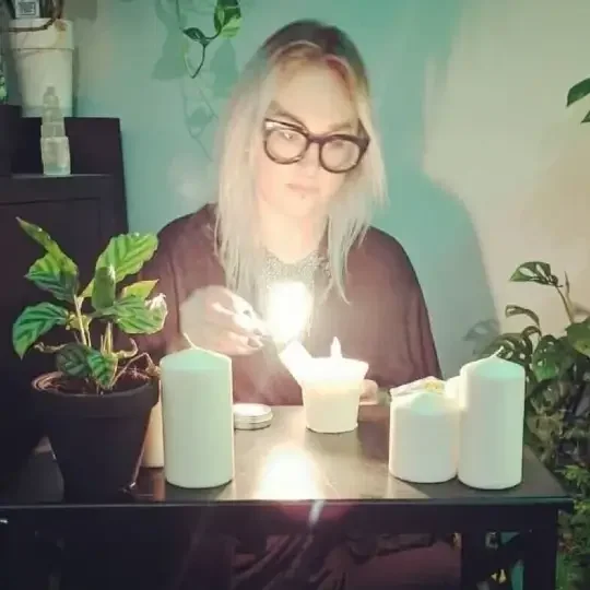 Maggie sits at a table with a lit match in her hand and she is holding it towards a white pillar candle | The Witch's Therapist | holistic psychotherapy in London Ontario