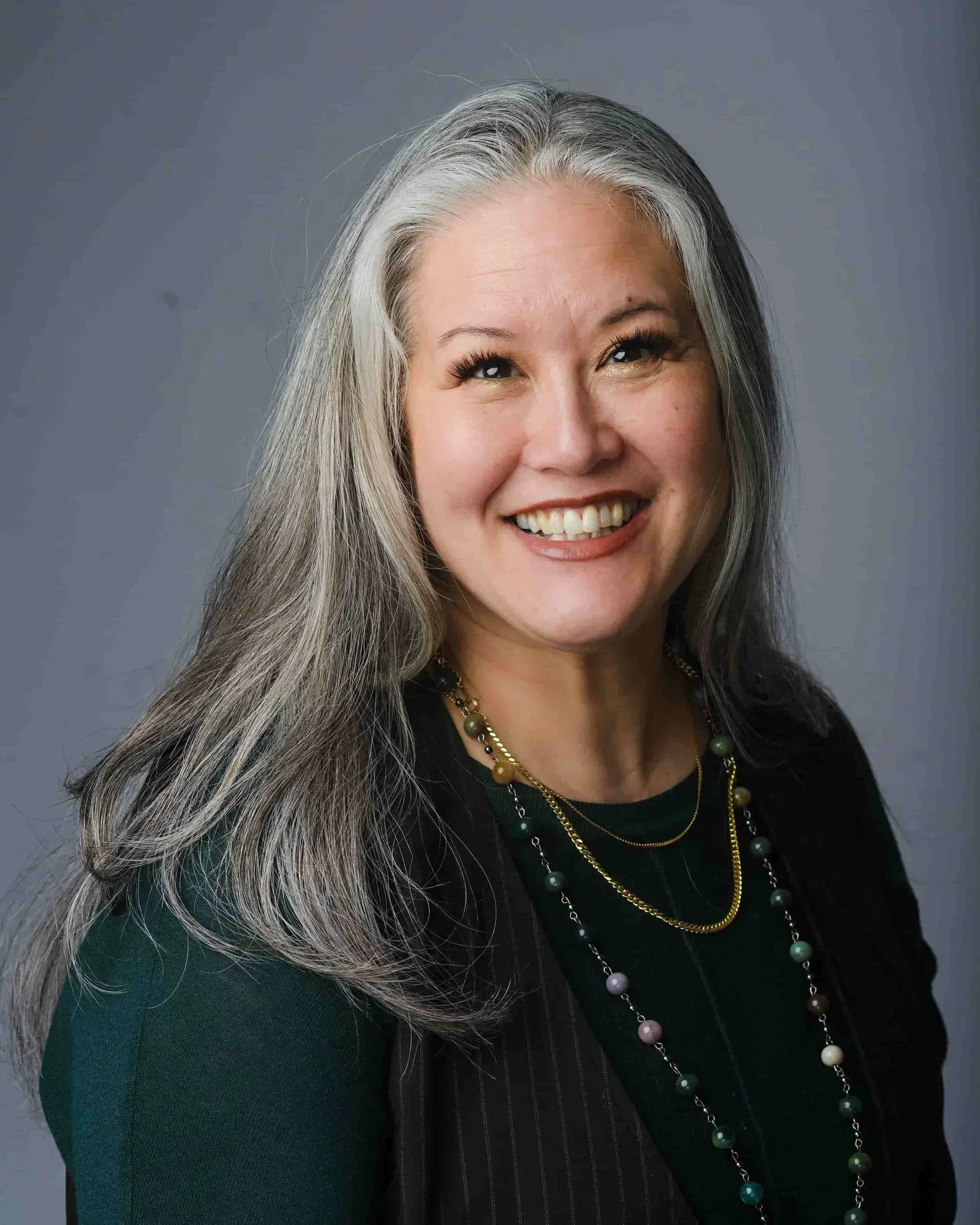 A woman with long gray hair, smiling, wearing a dark green top, a black jacket, and layered necklaces with beads of various colors.