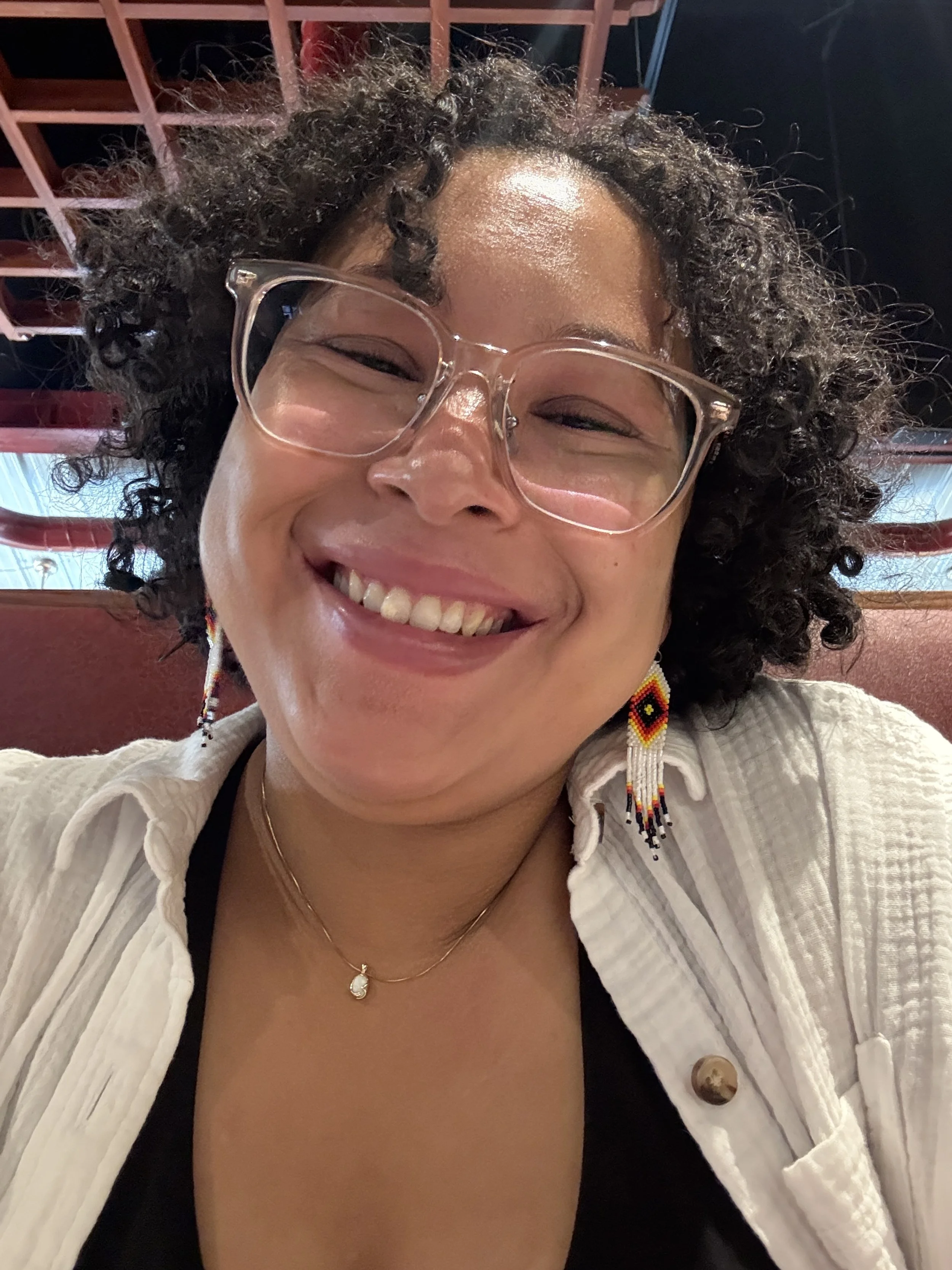 A woman smiling with curly hair, wearing glasses, colorful beaded earrings, a necklace, and a white shirt, sitting indoors with wooden and dark blue decor behind her.