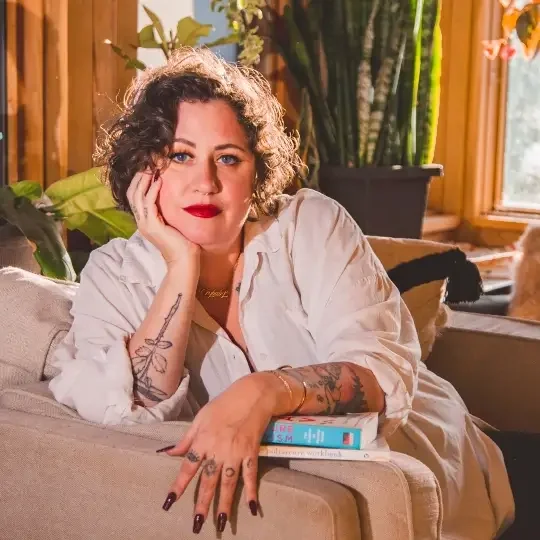 A woman with curly hair sitting on a beige sofa in a sunlit room surrounded by large green plants and orange flowers in pots. | The Witch's Therapist | holistic psychotherapy in London Ontario